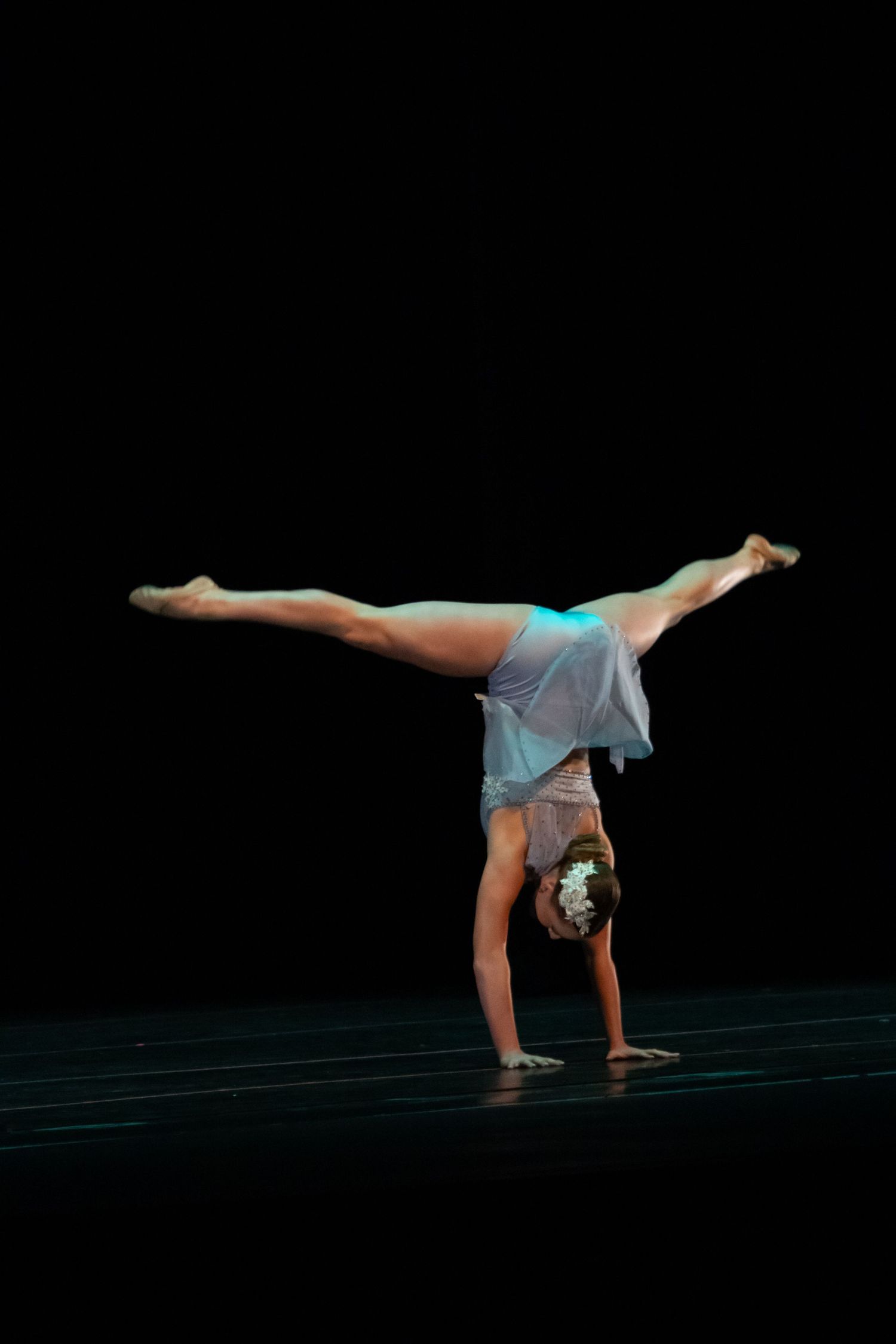 A dancer in a light blue dress performs a handstand split on a dark stage under teal lighting.