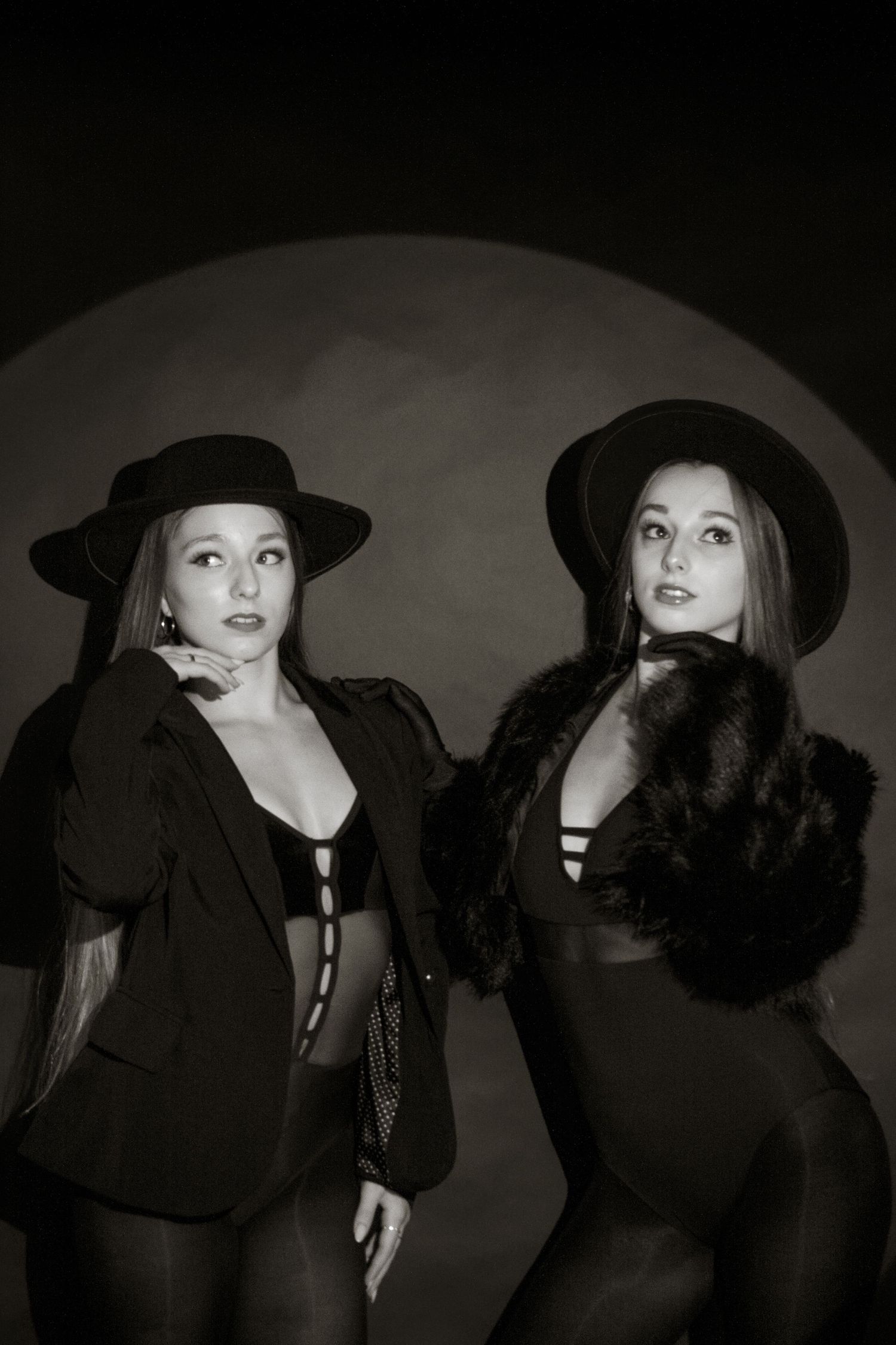 Two women in black hats and fur accessories pose together in dramatic black and white studio portrait.