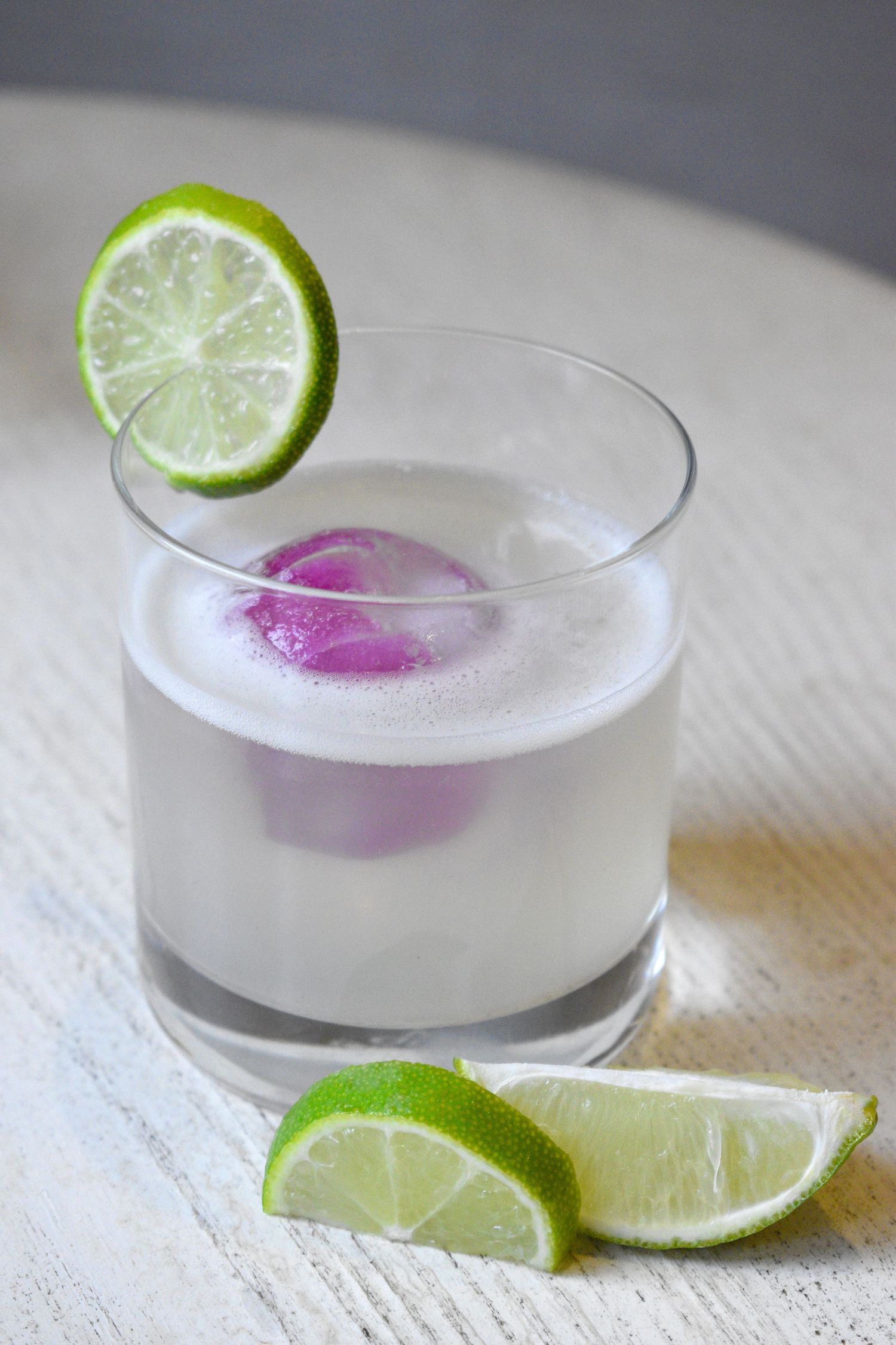 Frothy white lime cocktail in a rocks glass with purple ice spheres and fresh lime wheel garnish.