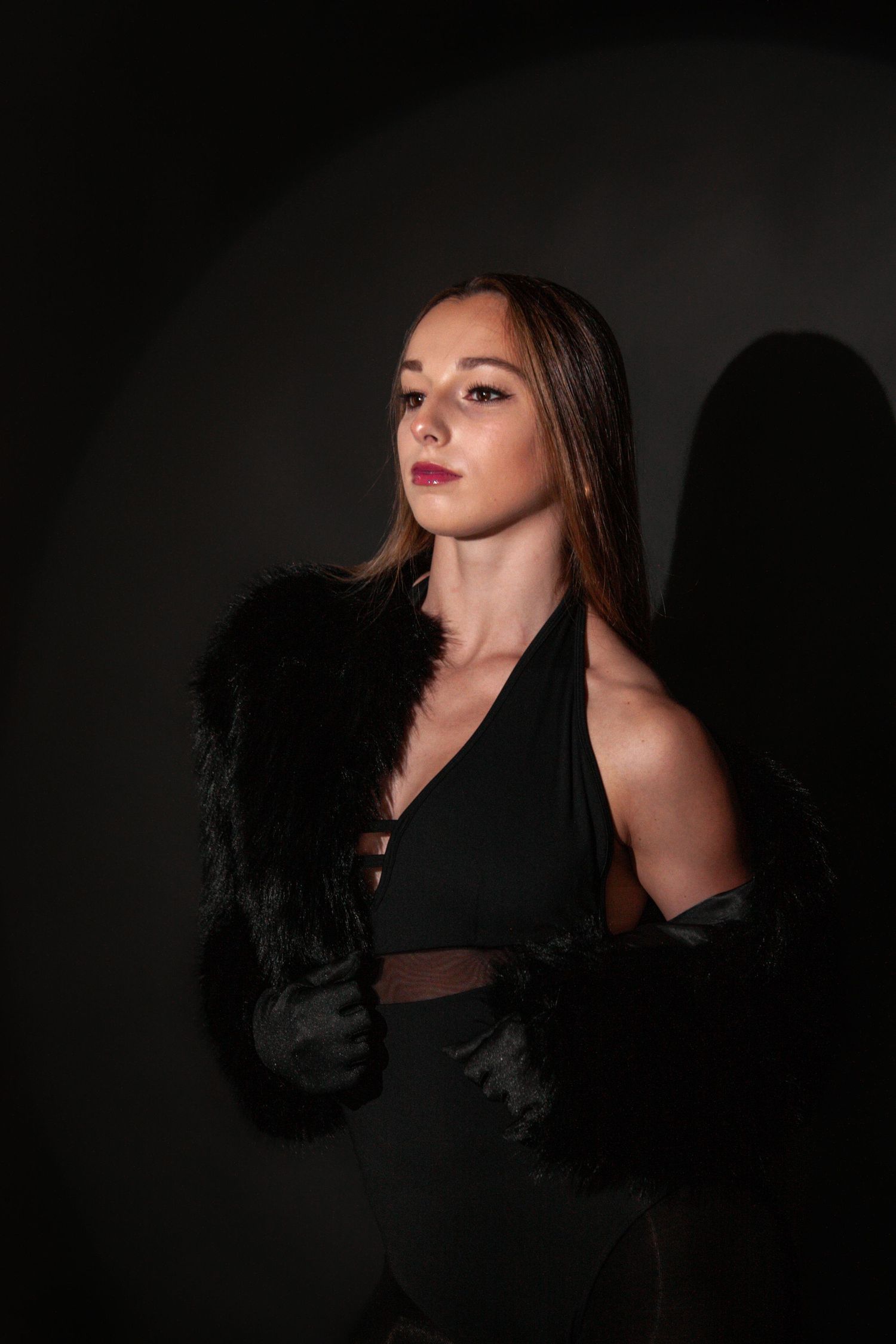 Young woman in black halter outfit with faux fur wrap poses dramatically against a dark studio background.