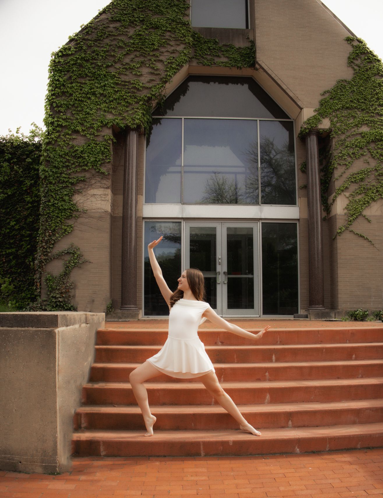 Ballet dancer in white dress poses on brick steps outside an ivy-covered building on pointe.