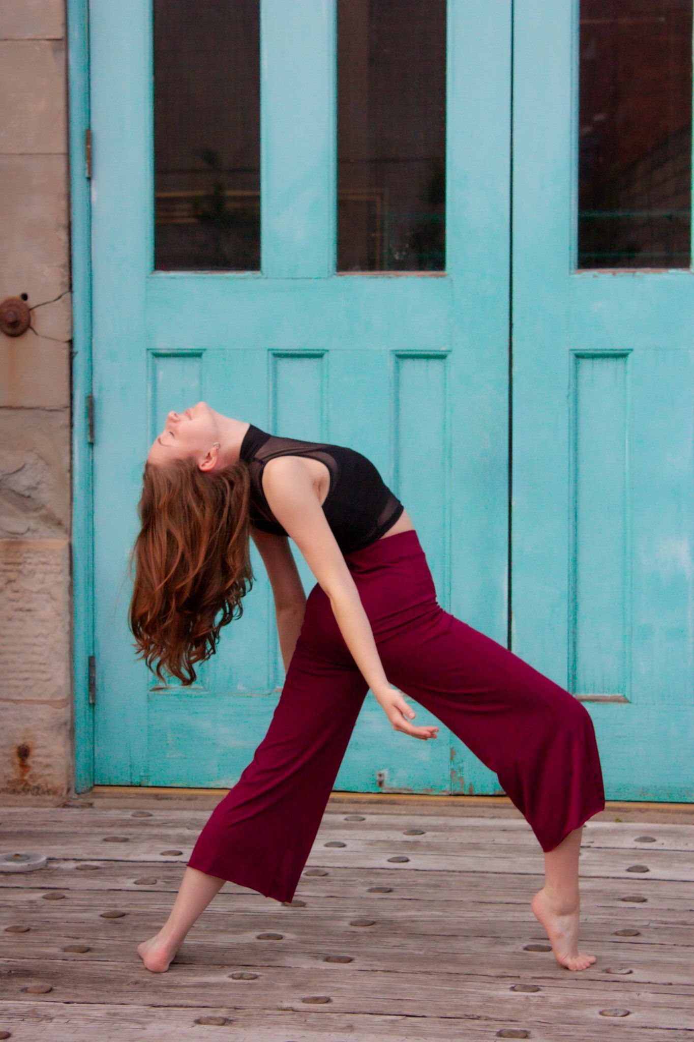 A contemporary dancer arches backward in burgundy wide-leg pants and black top before a turquoise wooden door.