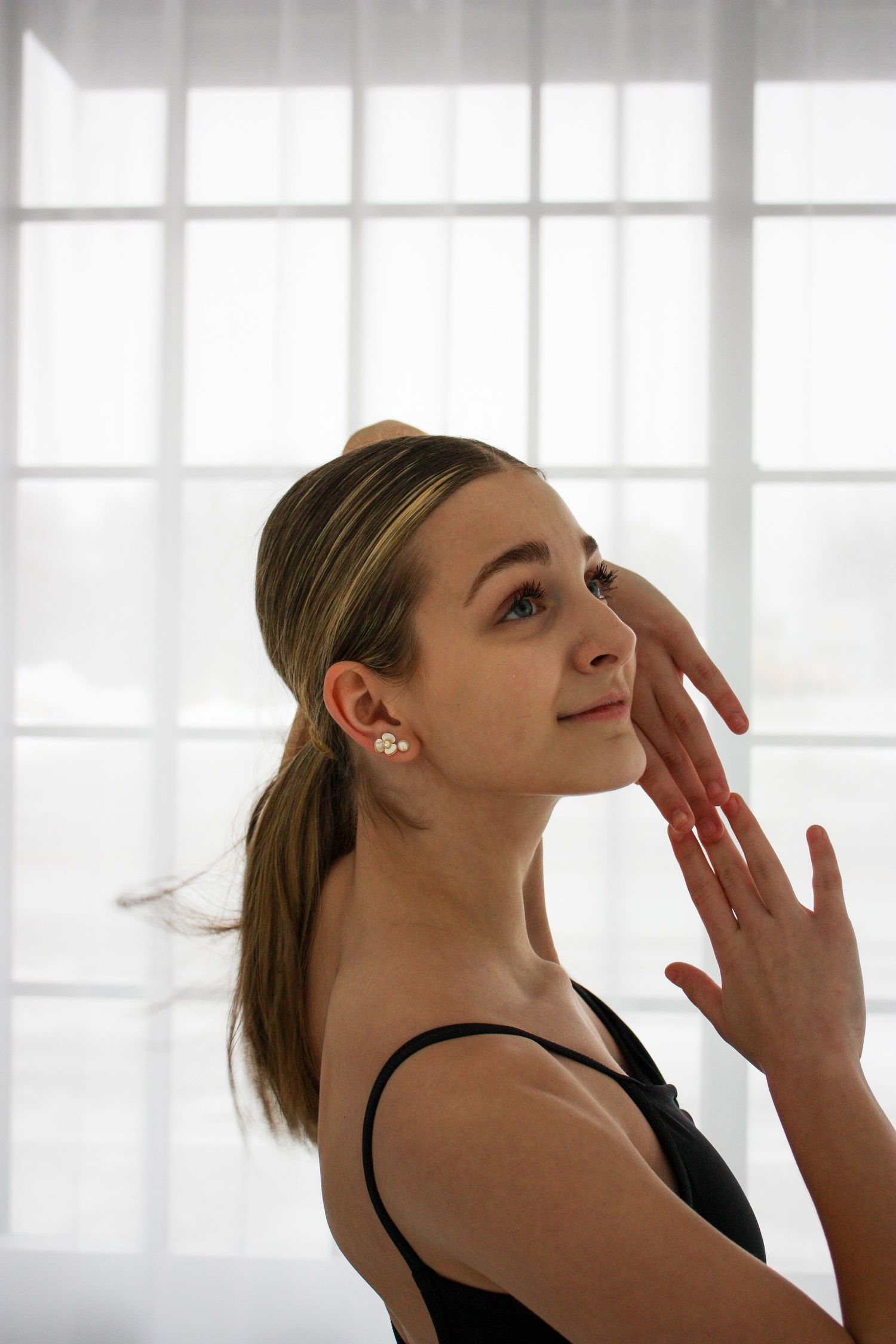Young female ballet dancer in black leotard posing gracefully with hand near face by bright window.