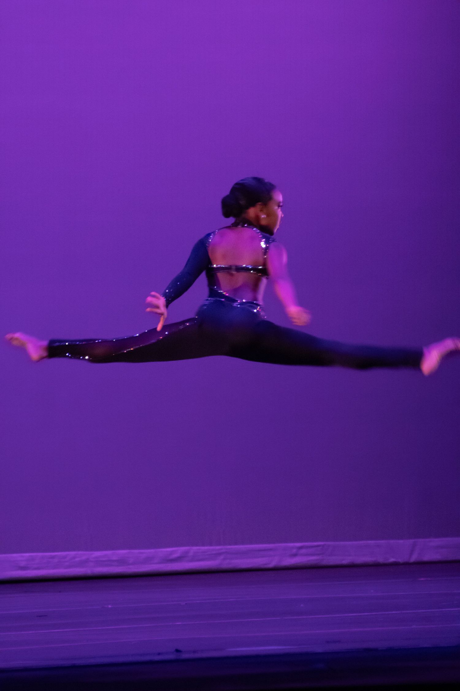 A dancer performs a mid-air split leap on stage in a sparkly black costume against a purple backdrop.