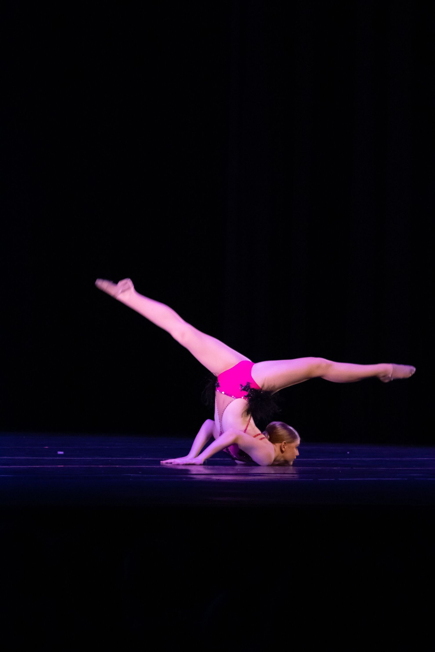 A dancer in a pink leotard performs an acrobatic move on stage.
