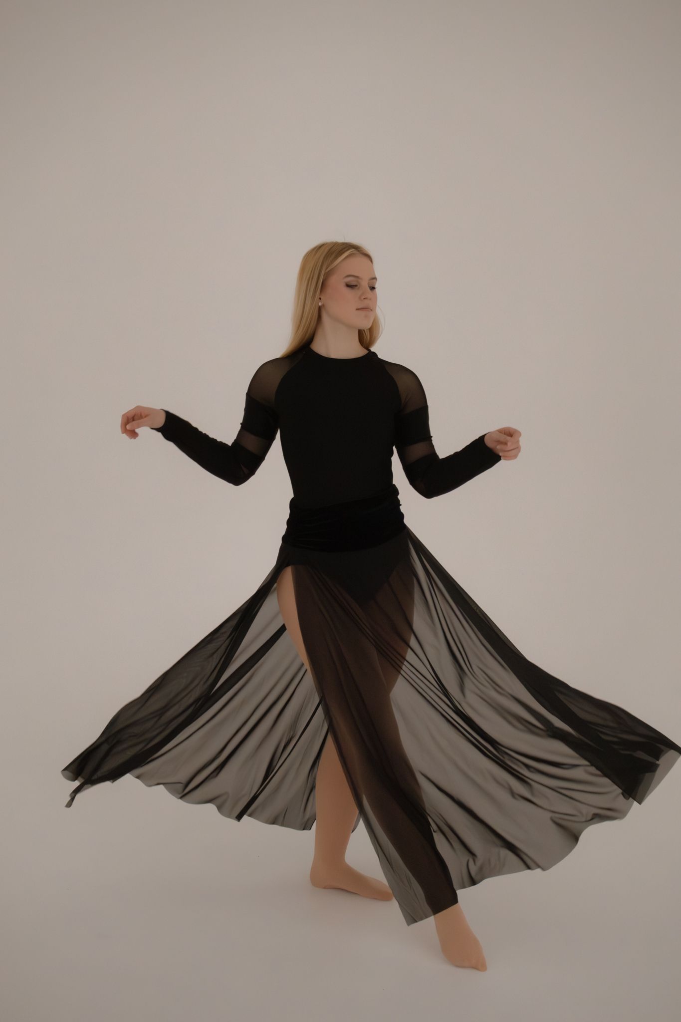 Blonde dancer in black long-sleeve bodysuit and flowing sheer skirt twirls on white background.