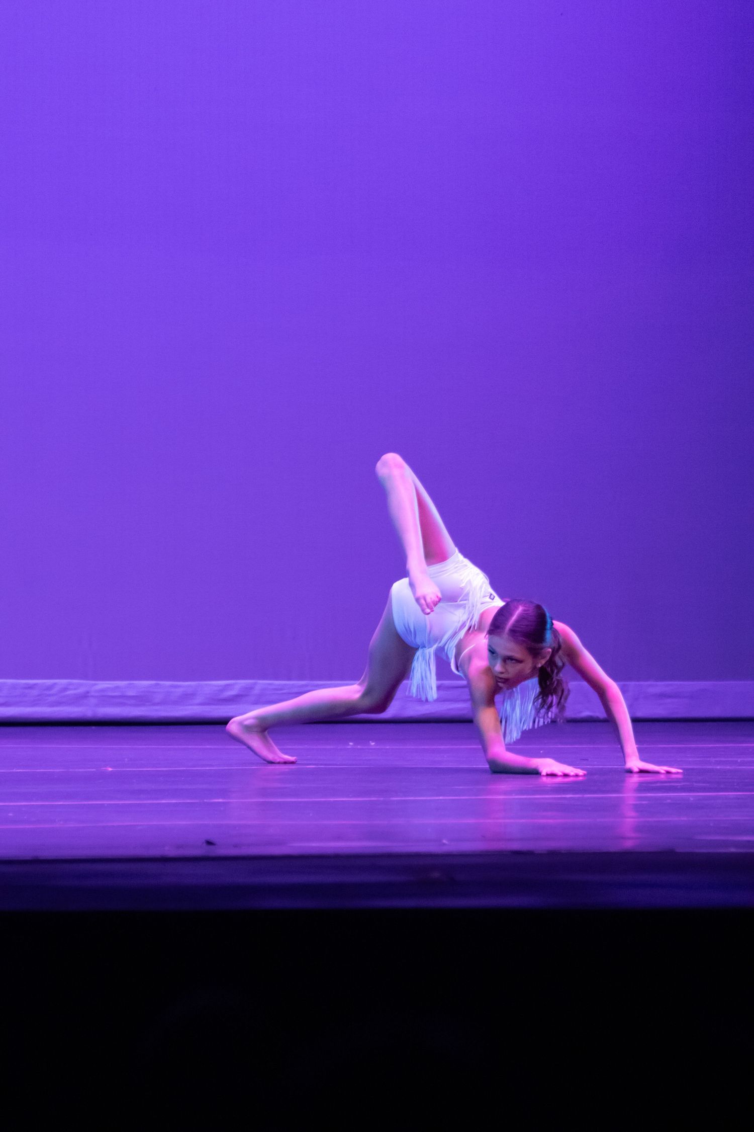 A dancer in a white fringed costume performs a low acrobatic floor move under purple stage lights.