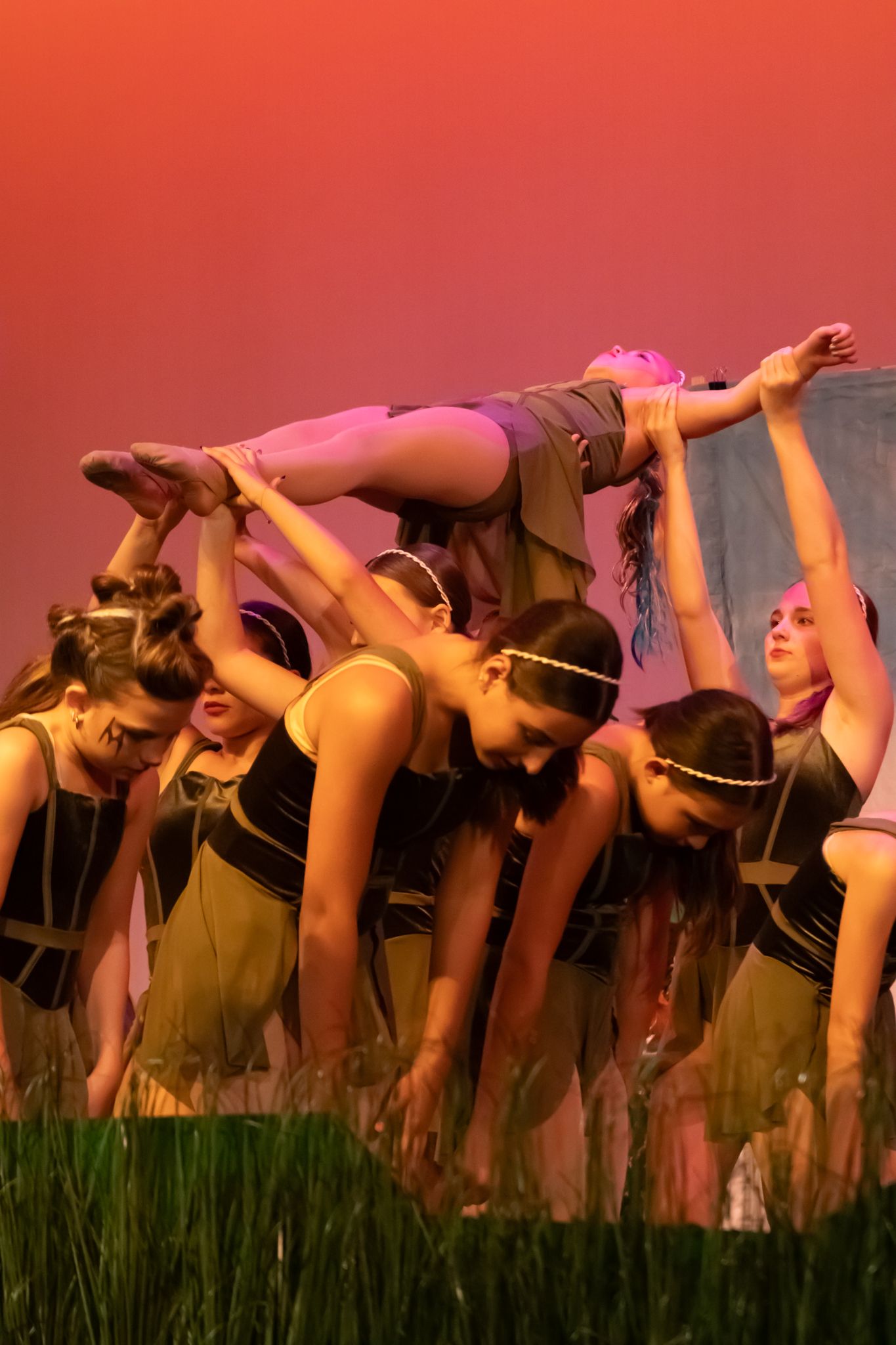 Group of female dancers lift a performer overhead in a dramatic stage production with warm orange lighting.