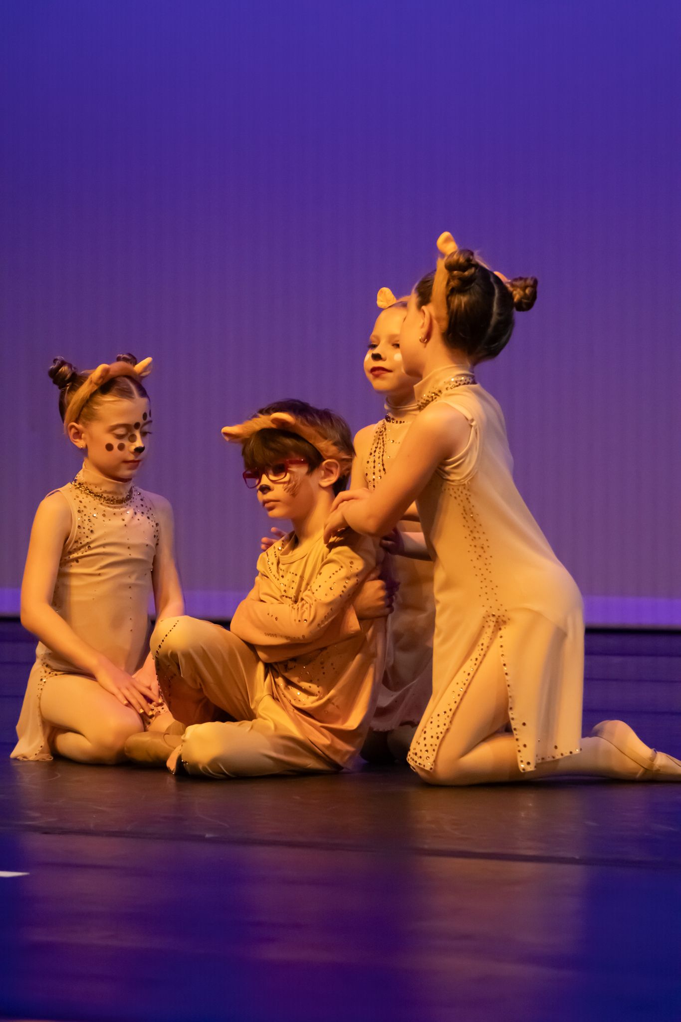 Four young dancers in cat costumes perform an expressive stage routine under purple stage lighting.