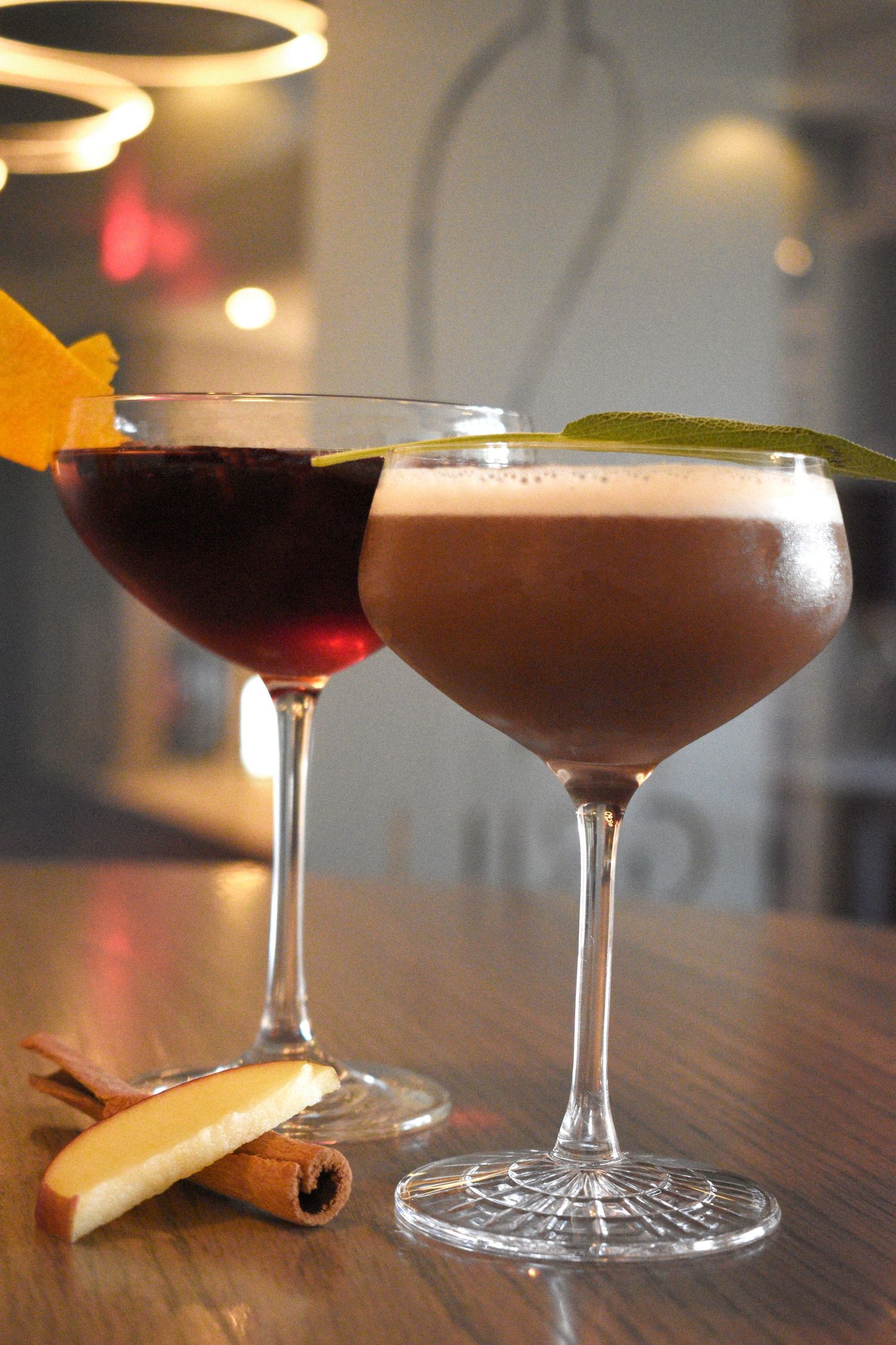 Two elegant cocktails in coupe glasses garnished with orange peel and sage on a bar.