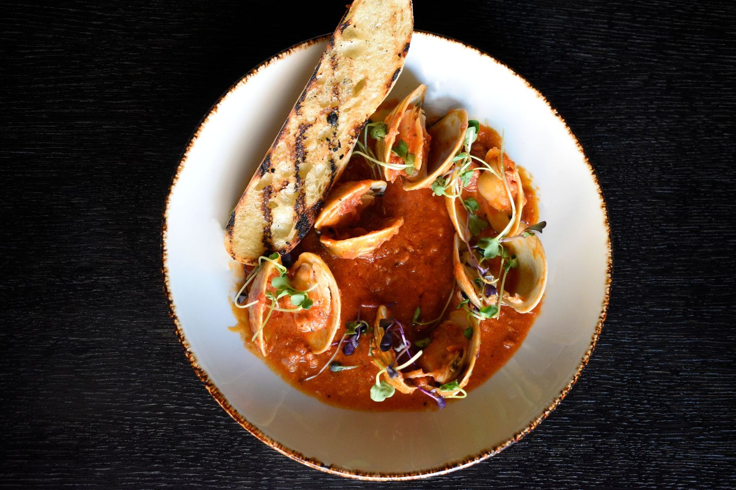 Overhead view of a rich seafood stew with clams, crab, and grilled bread in a white bowl.