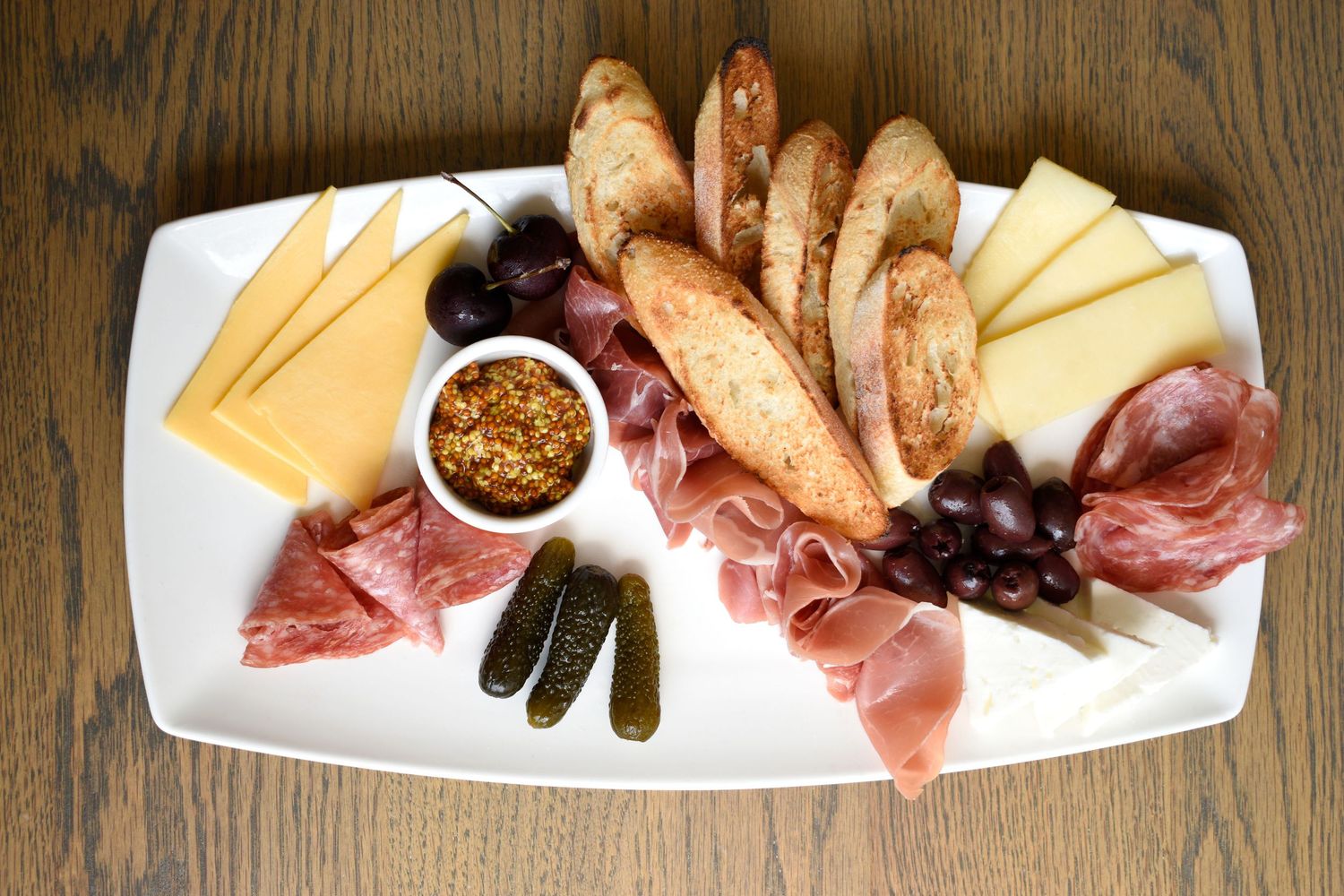Charcuterie board with prosciutto, salami, cheese, baguette slices, grapes, pickles, and whole grain mustard.