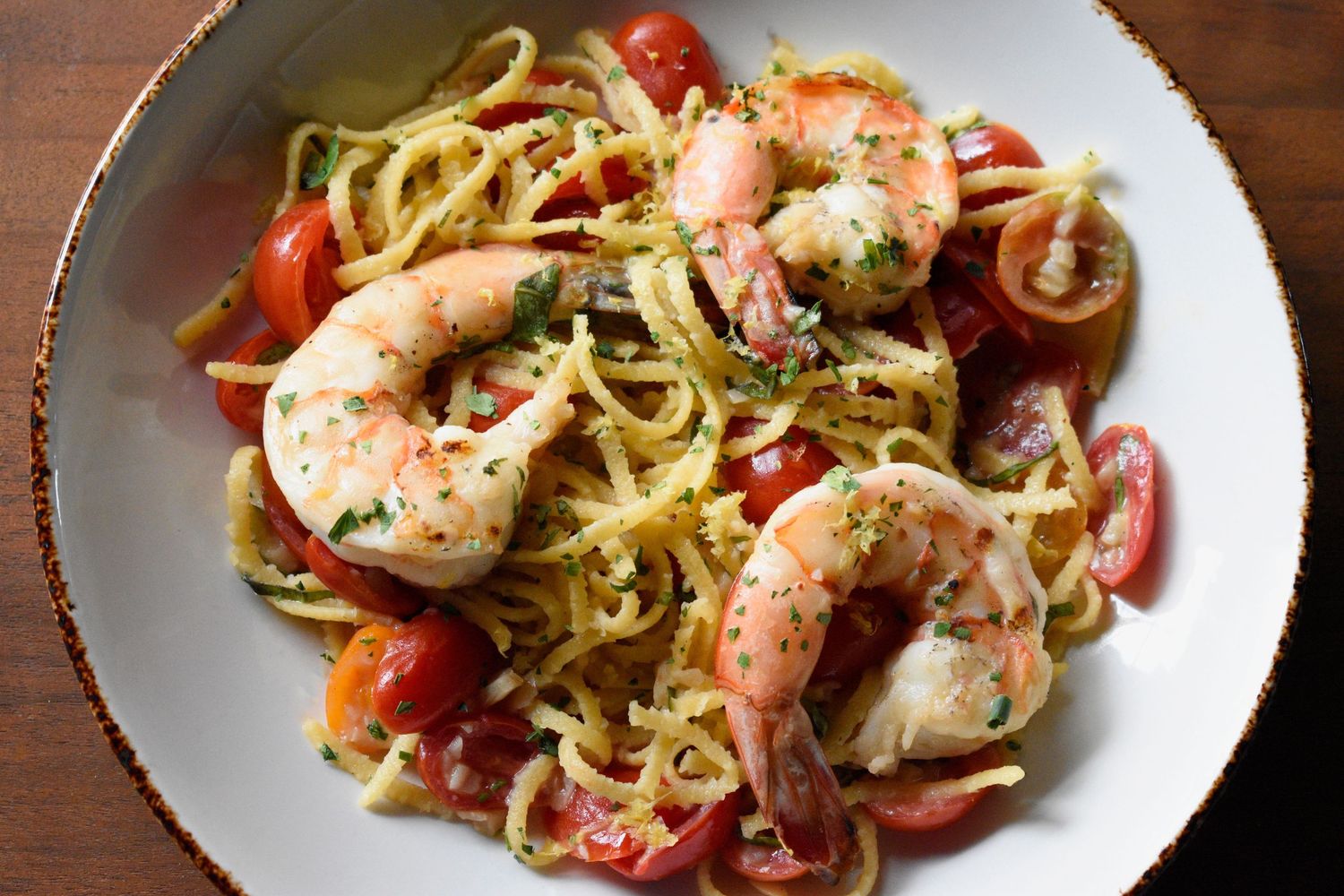 Shrimp scampi pasta with cherry tomatoes and herbs in a white bowl on a wooden table.