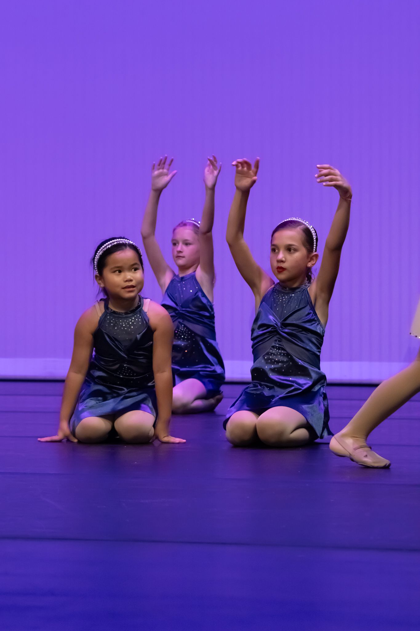 Young girls in blue dresses kneel on stage with arms raised during a dance recital performance.