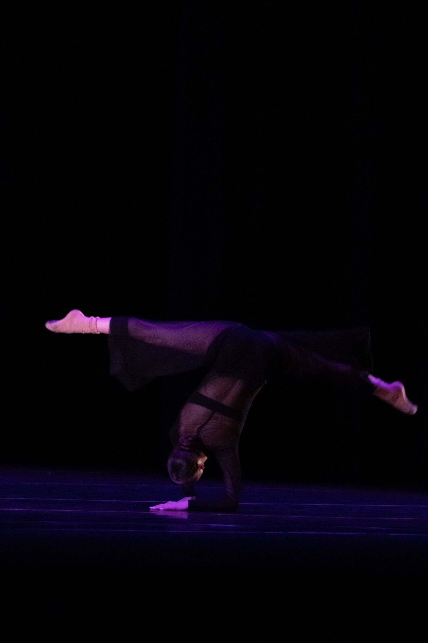 Dancer in black performs a headstand split silhouetted against a dark stage backdrop.