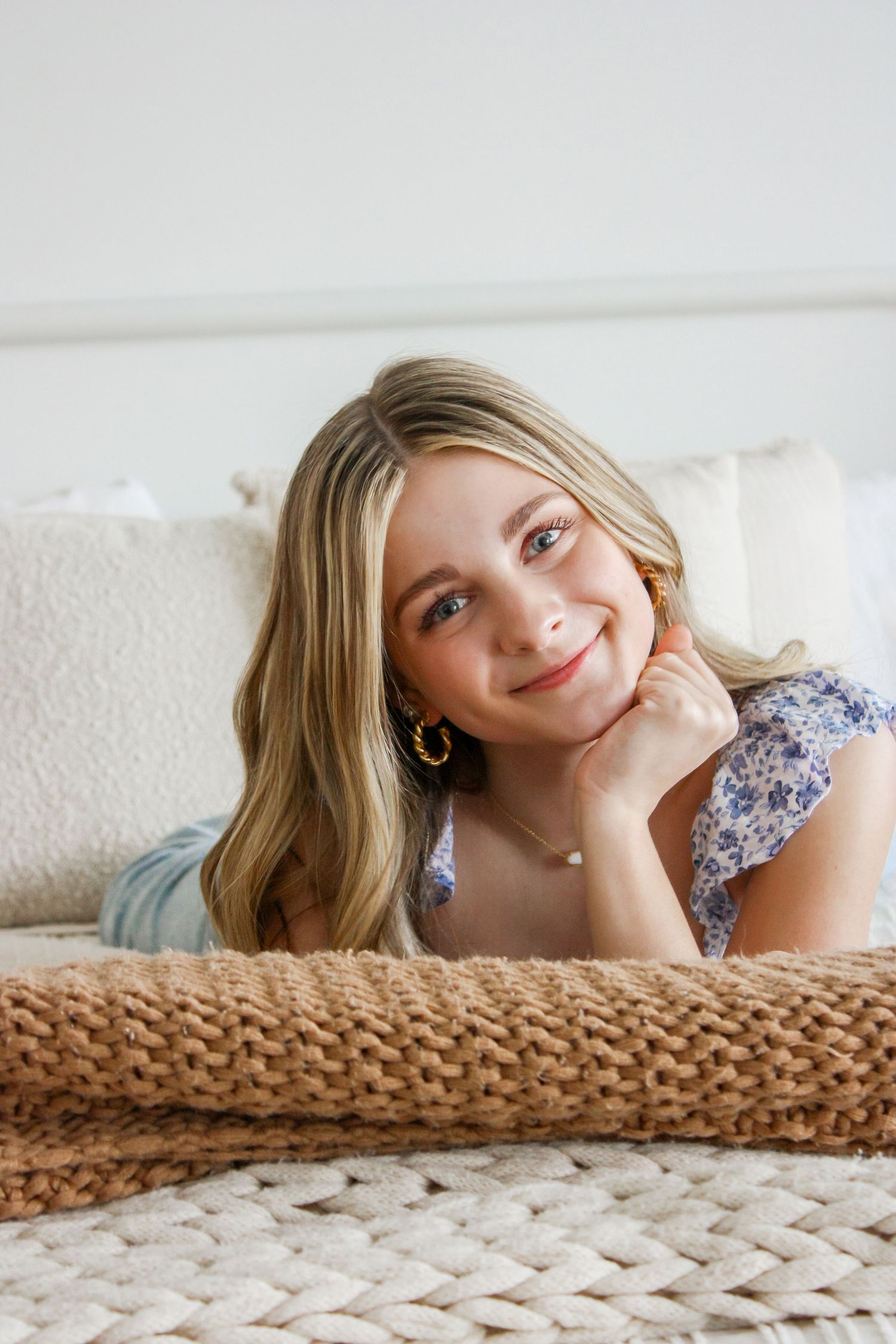 Smiling blonde woman in floral top lying on bed, resting chin on hand, wearing gold earrings.