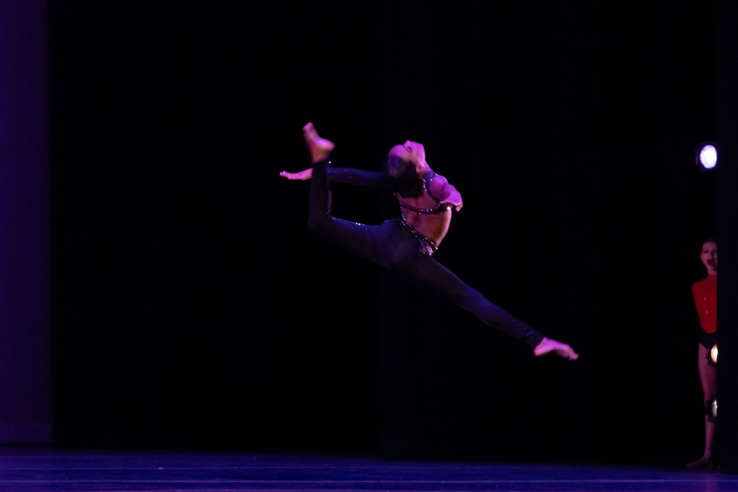 A dancer leaps dramatically in mid-air against a dark stage background with another performer visible behind.