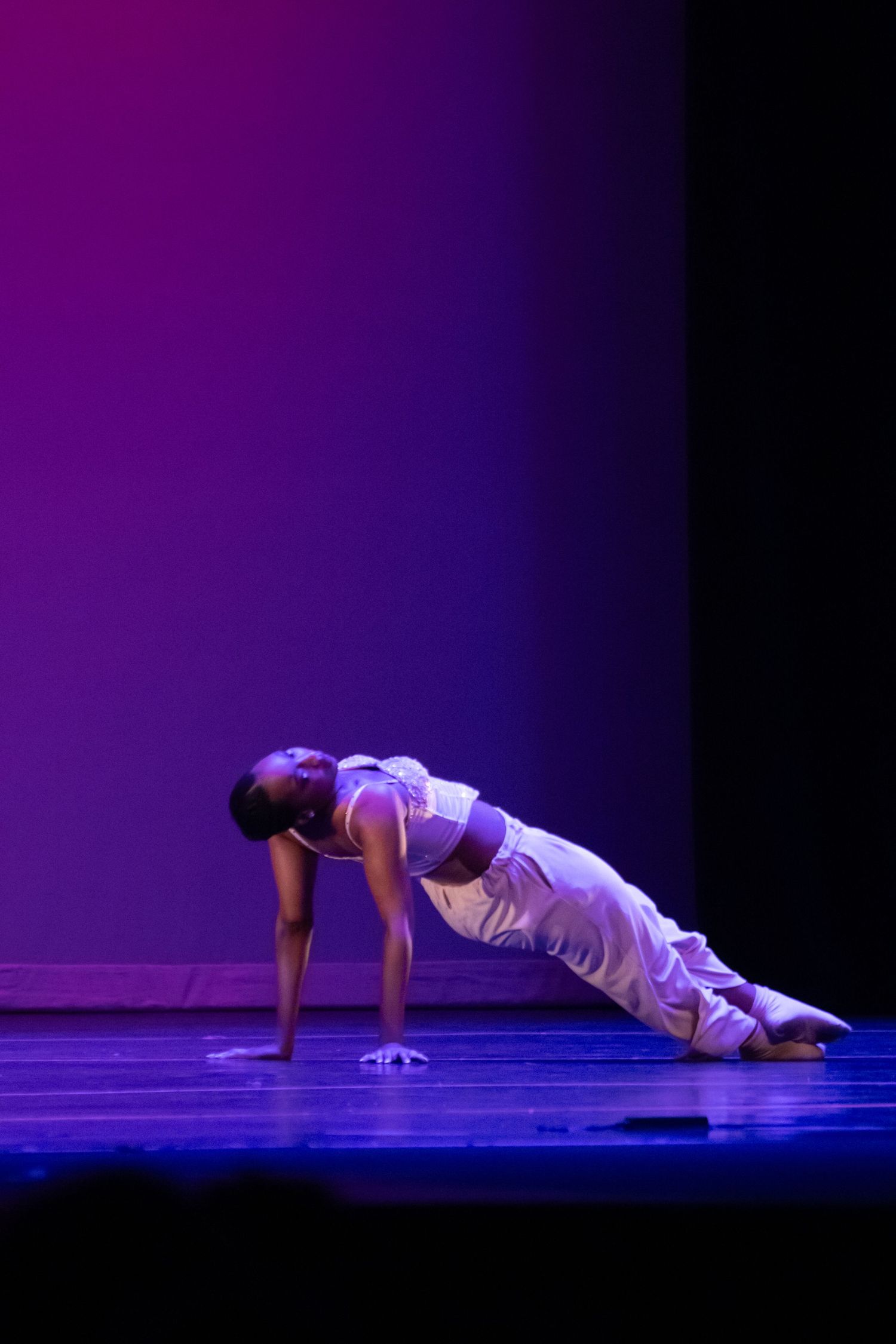A dancer in white performs a dramatic floor pose on a purple-lit recital stage.