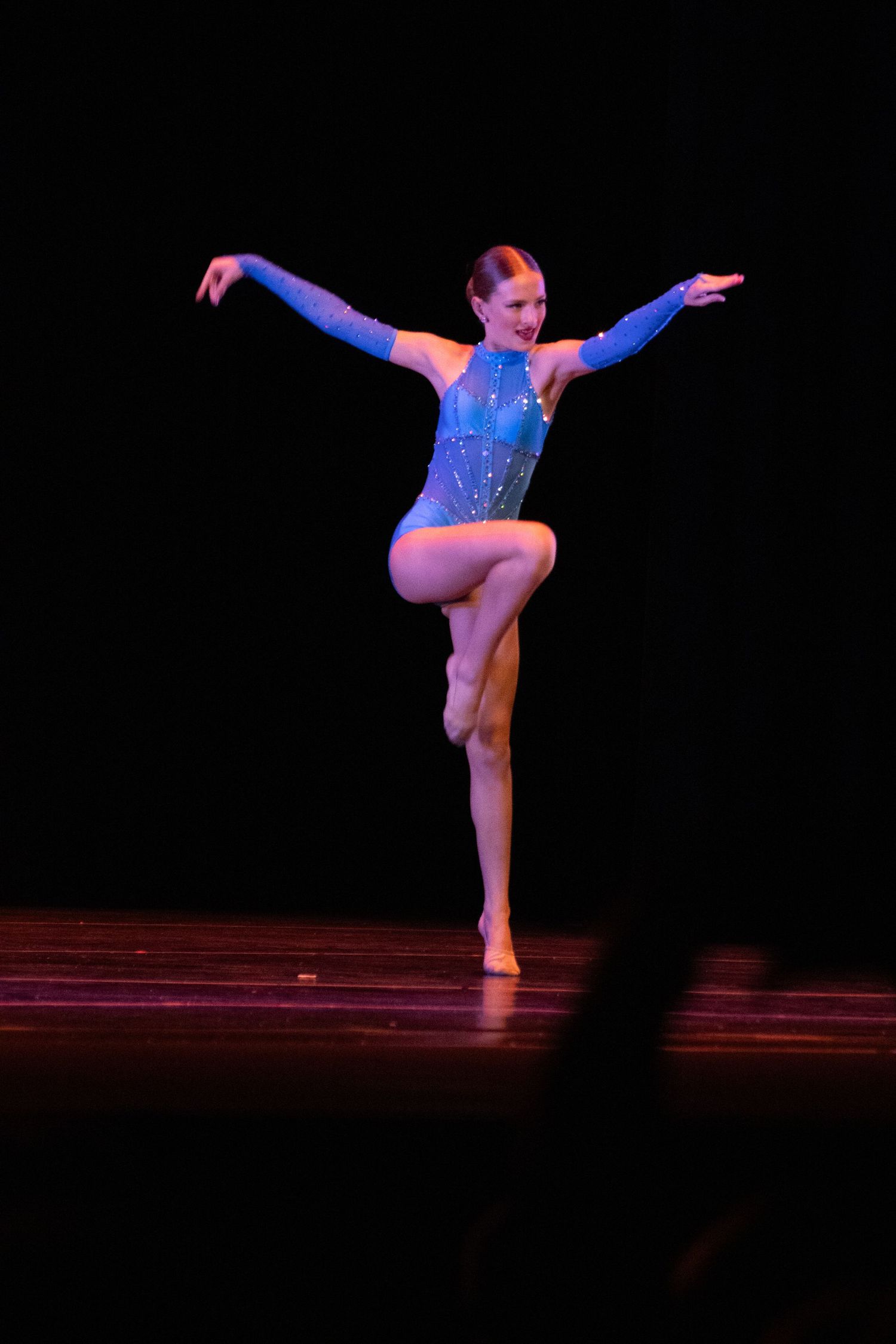 Girl in blue sequined leotard balances on one leg with arms extended during a dance performance.