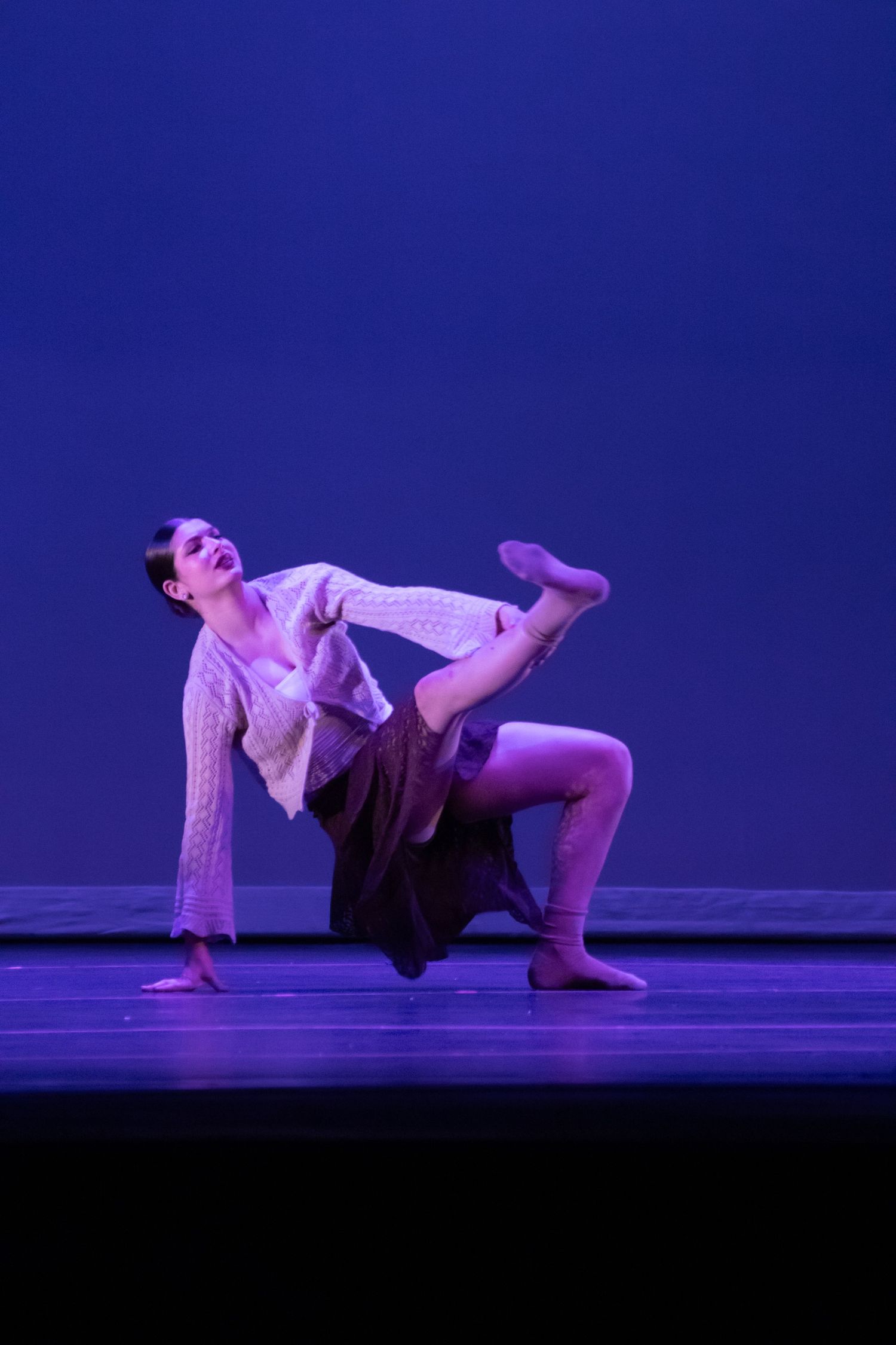 A contemporary dancer in flowing costume performs an expressive low crouching movement on a purple-lit stage.