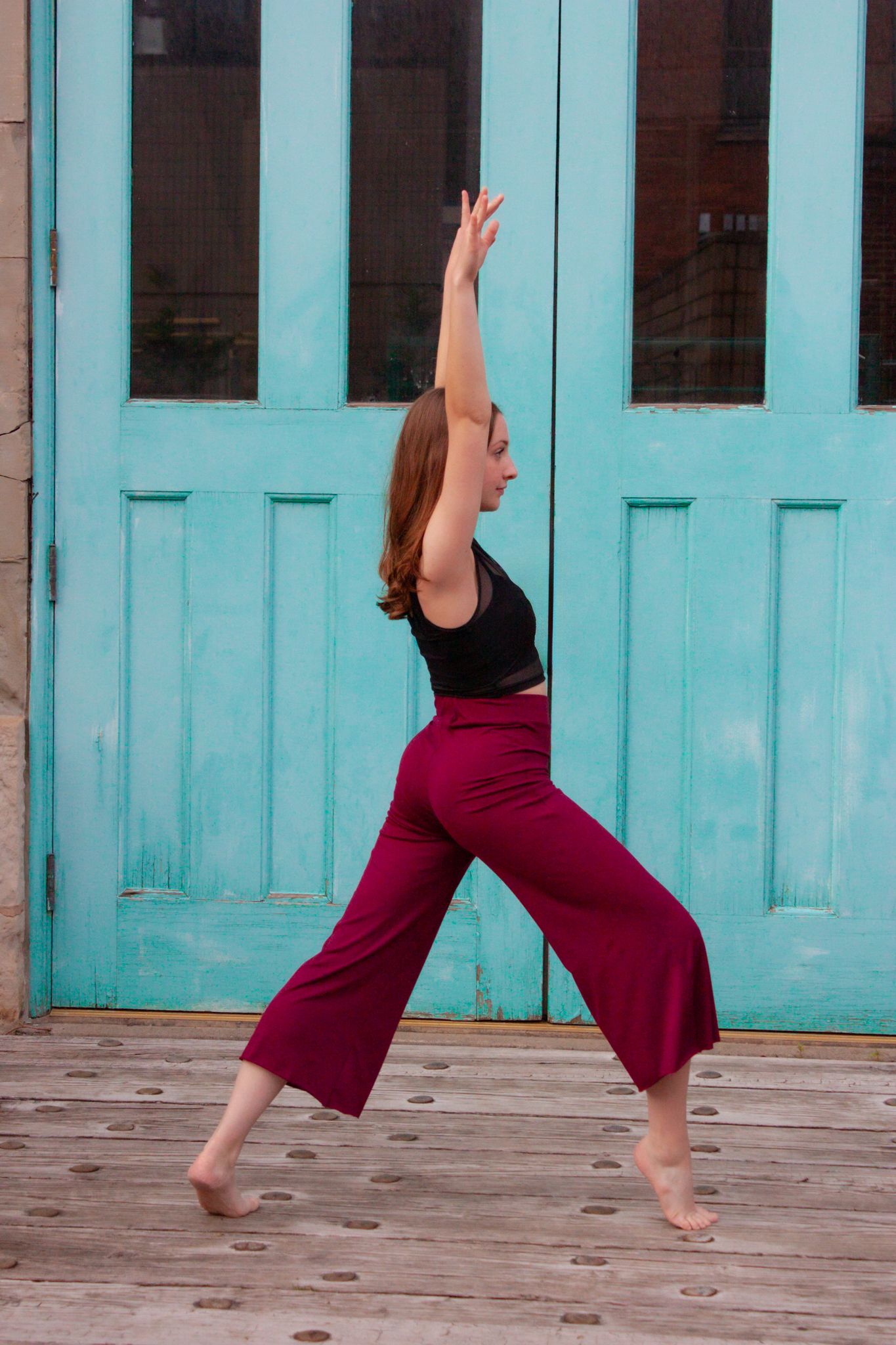 A dancer in a black top and magenta wide-leg pants performs a graceful pose in front of teal double doors.