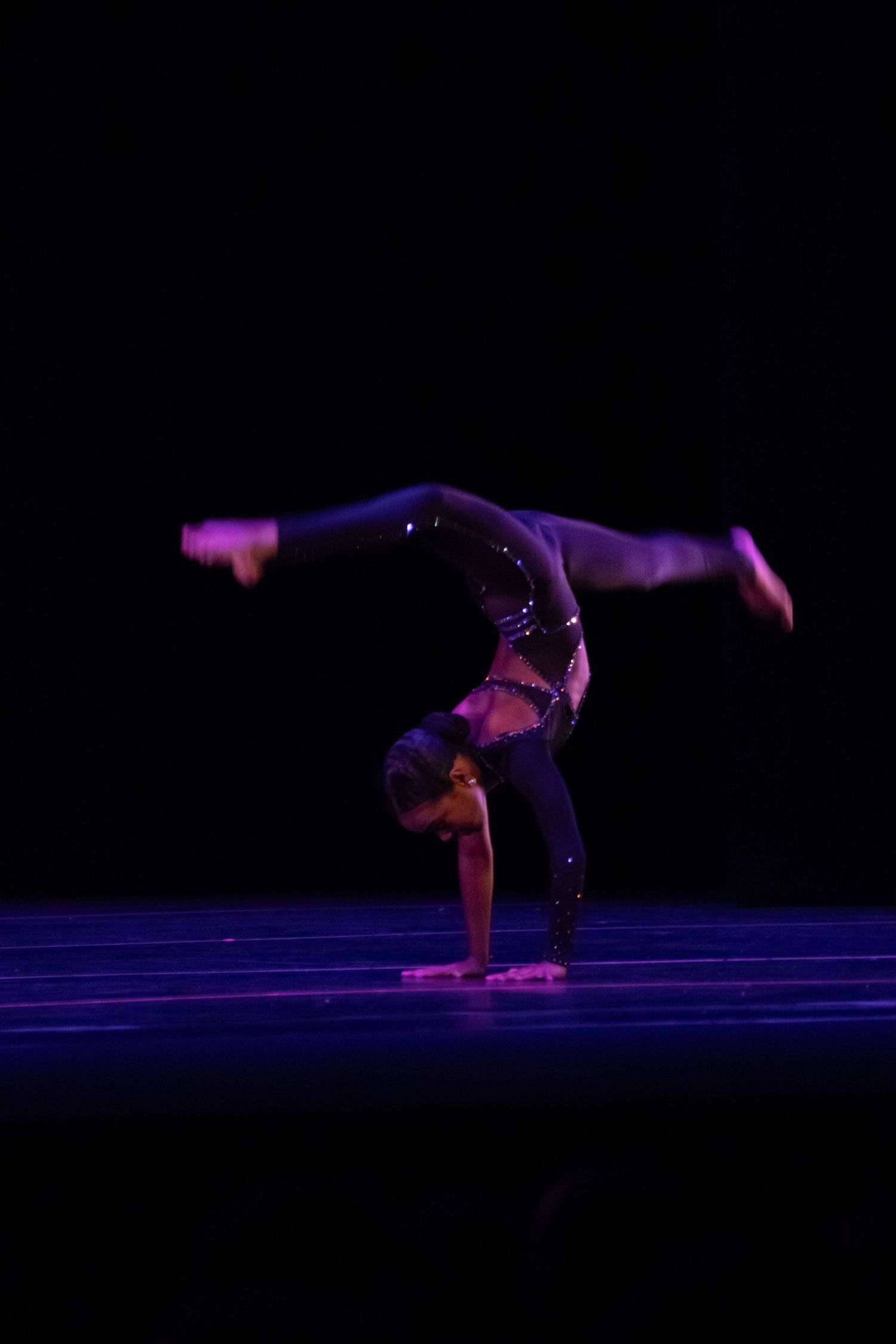 A dancer executes a handstand with legs split on a darkened stage under purple lighting.