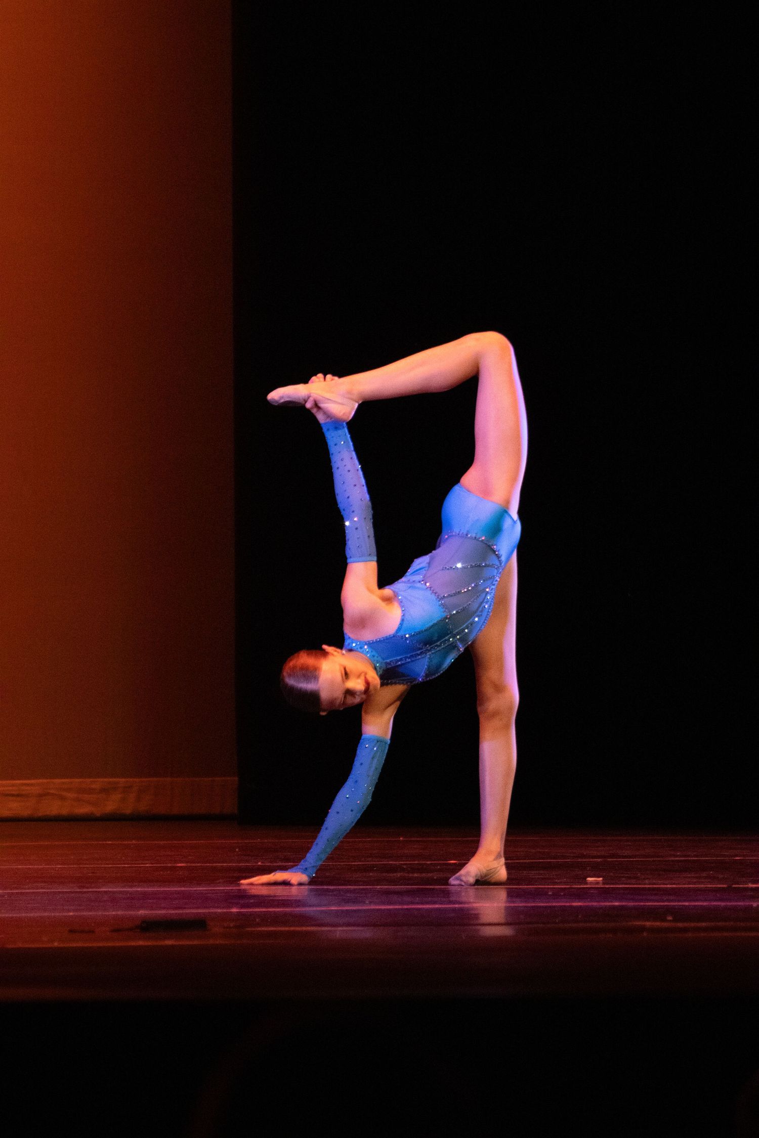 Young gymnast in blue sequined leotard performs impressive backbend on stage during dance recital.