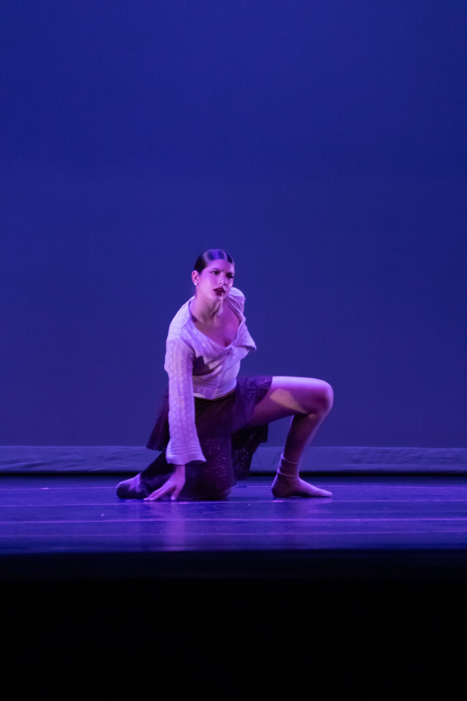 A contemporary dancer in a white top and dark skirt performs a dramatic kneeling pose on a blue-lit stage.