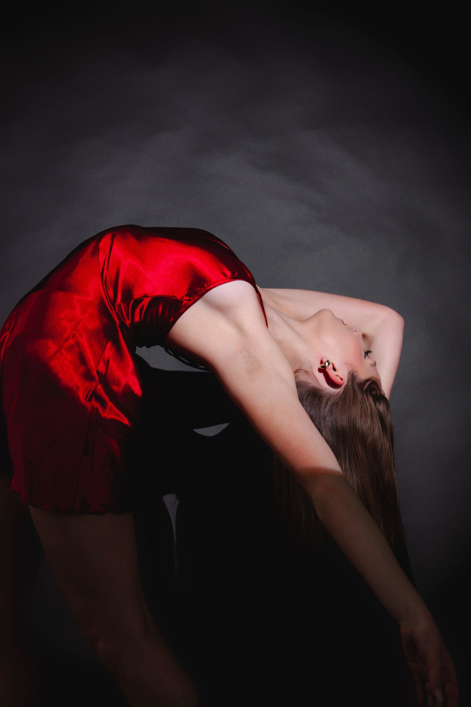 Dancer in red satin dress performs dramatic backbend with flowing hair against dark background.