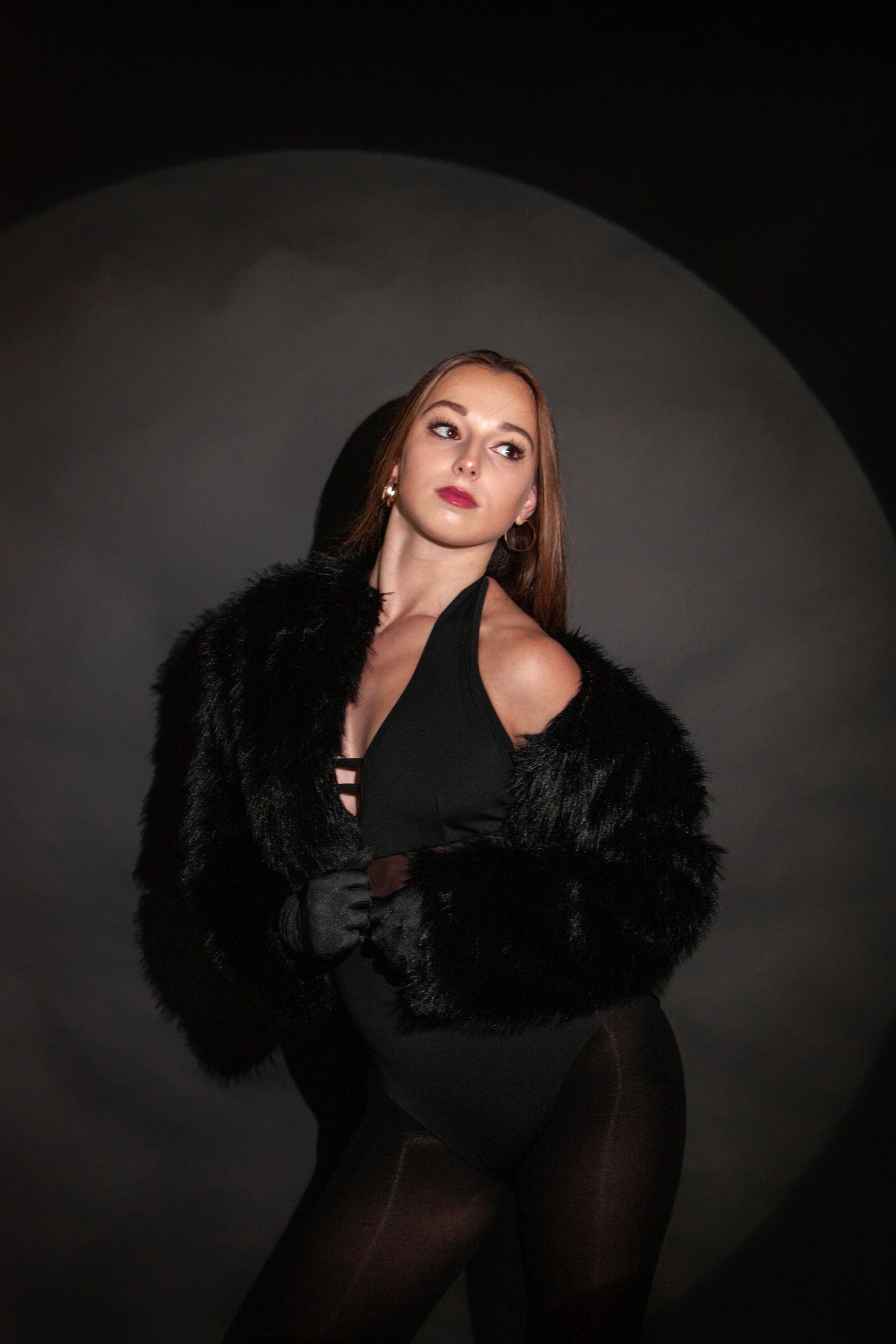 Woman in black fur coat and bodysuit poses dramatically against dark studio background with circular light.
