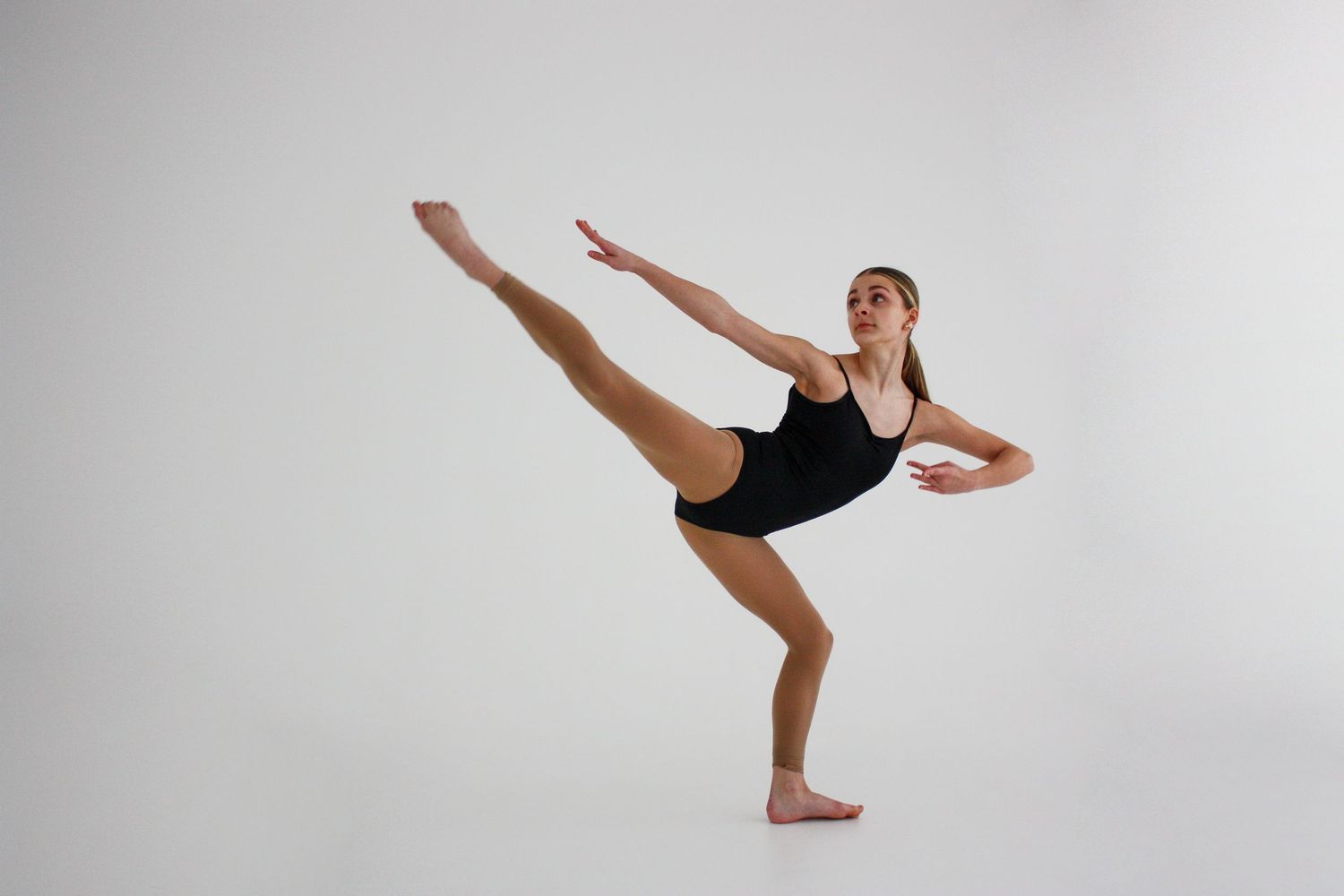 Young dancer in black leotard executing a high leg extension balance on one foot against white backdrop.