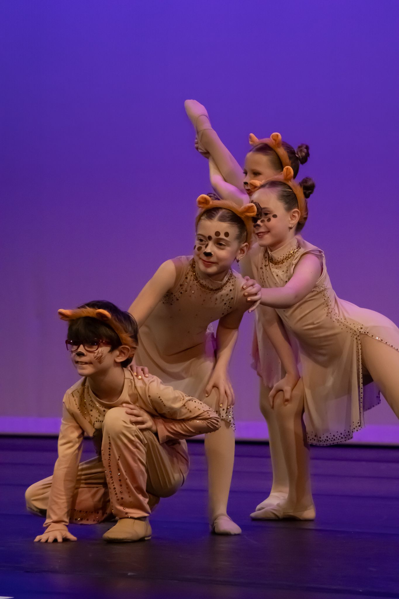 Three young children in lion costumes with face paint perform ballet on a purple-lit stage.