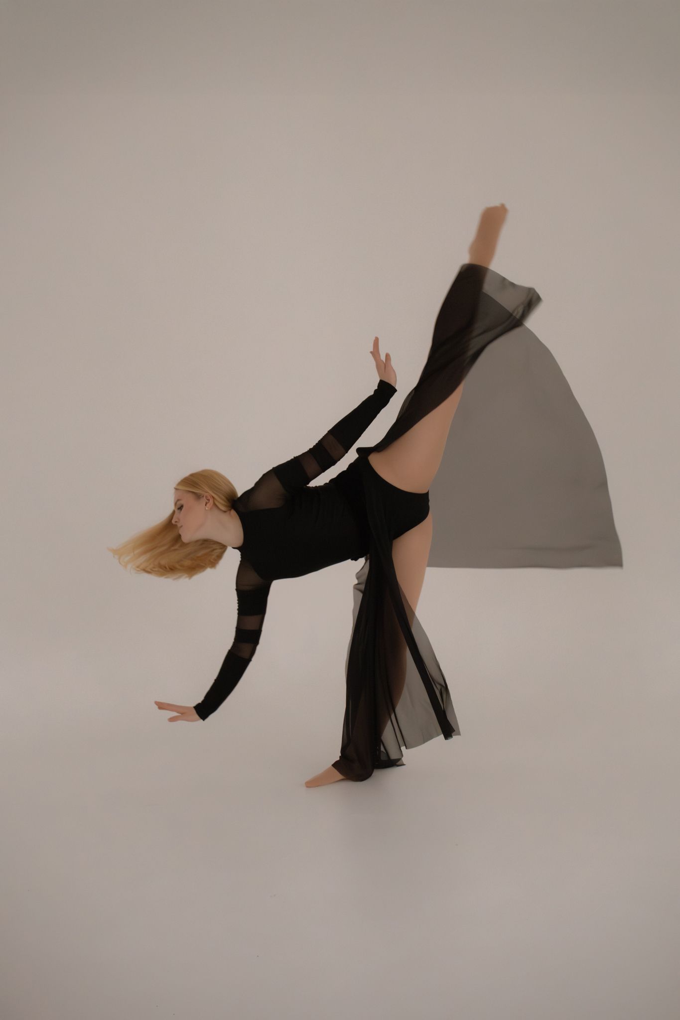 Blonde dancer in black bodysuit and sheer skirt performs a high leg extension on white background.