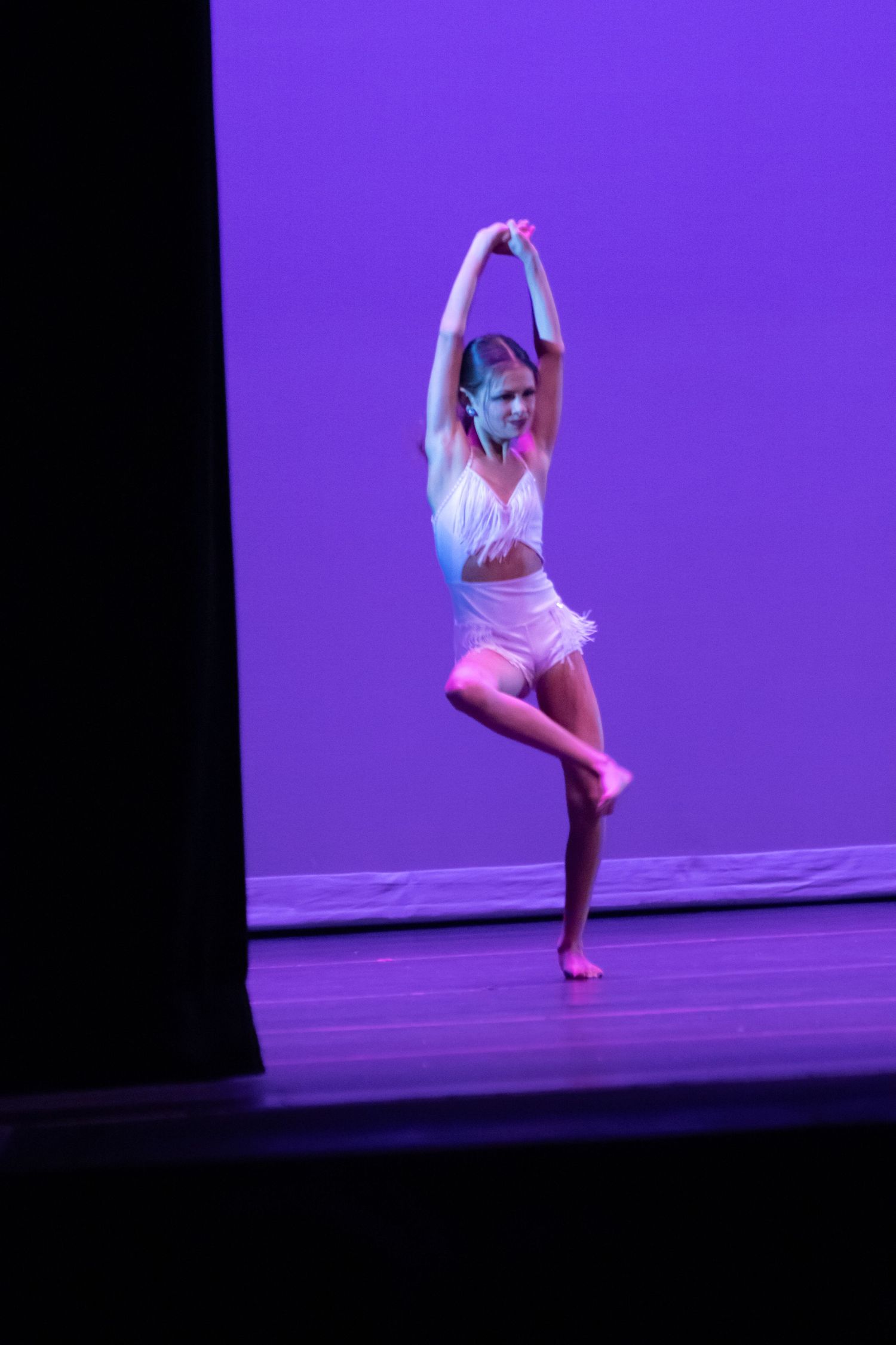 A young dancer in a white costume balances on one leg under purple stage lighting.