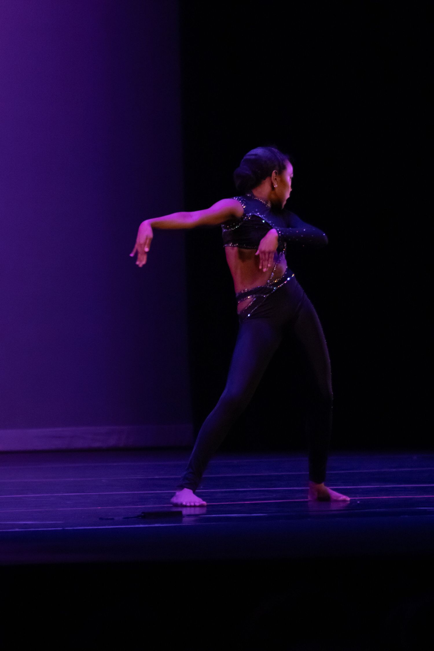 A dancer performs on a dark stage wearing a black outfit with a sequined top under purple lighting.