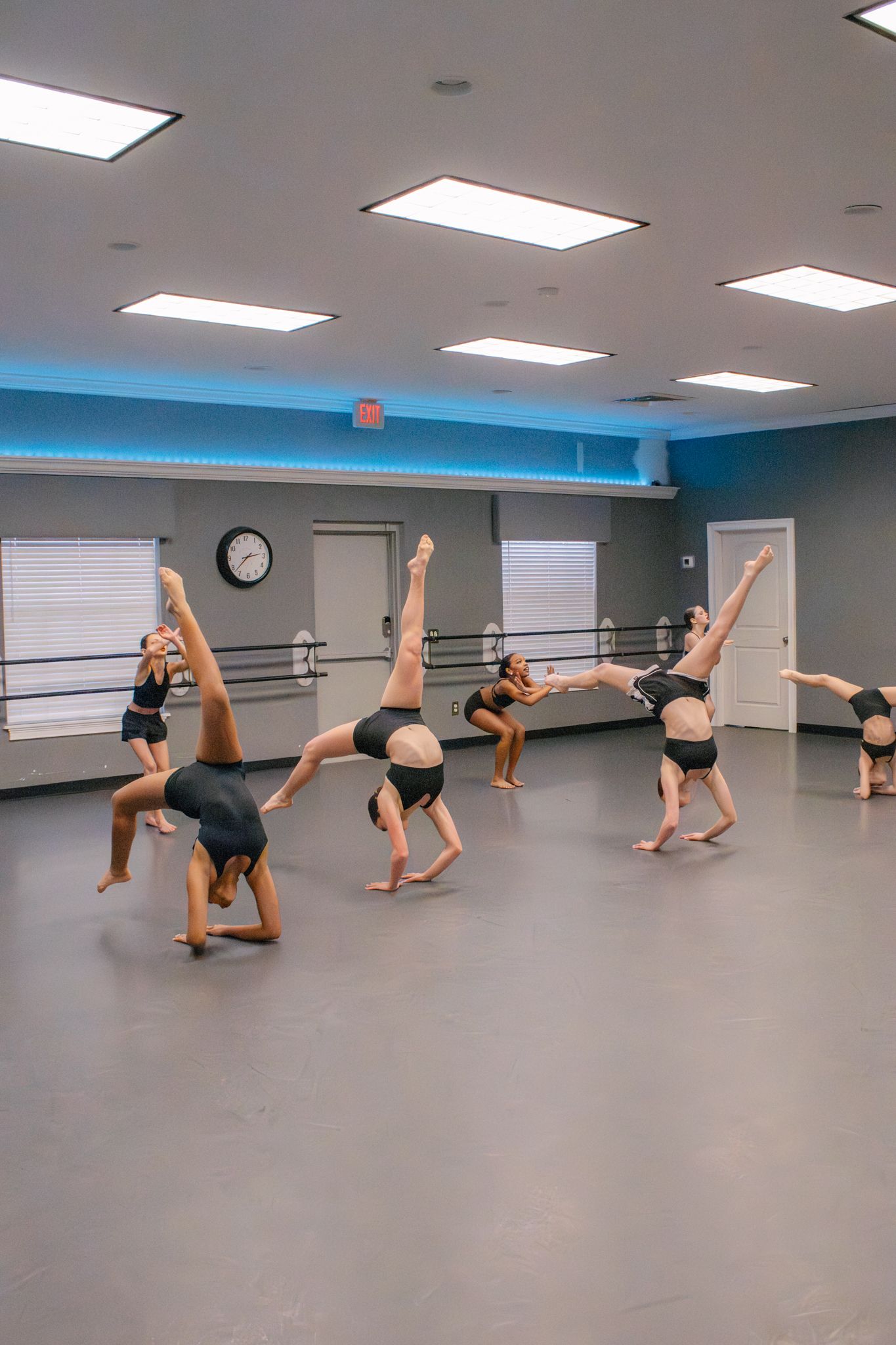 Dancers in black leotards perform acrobatic back walkovers in a bright studio with ballet barres.