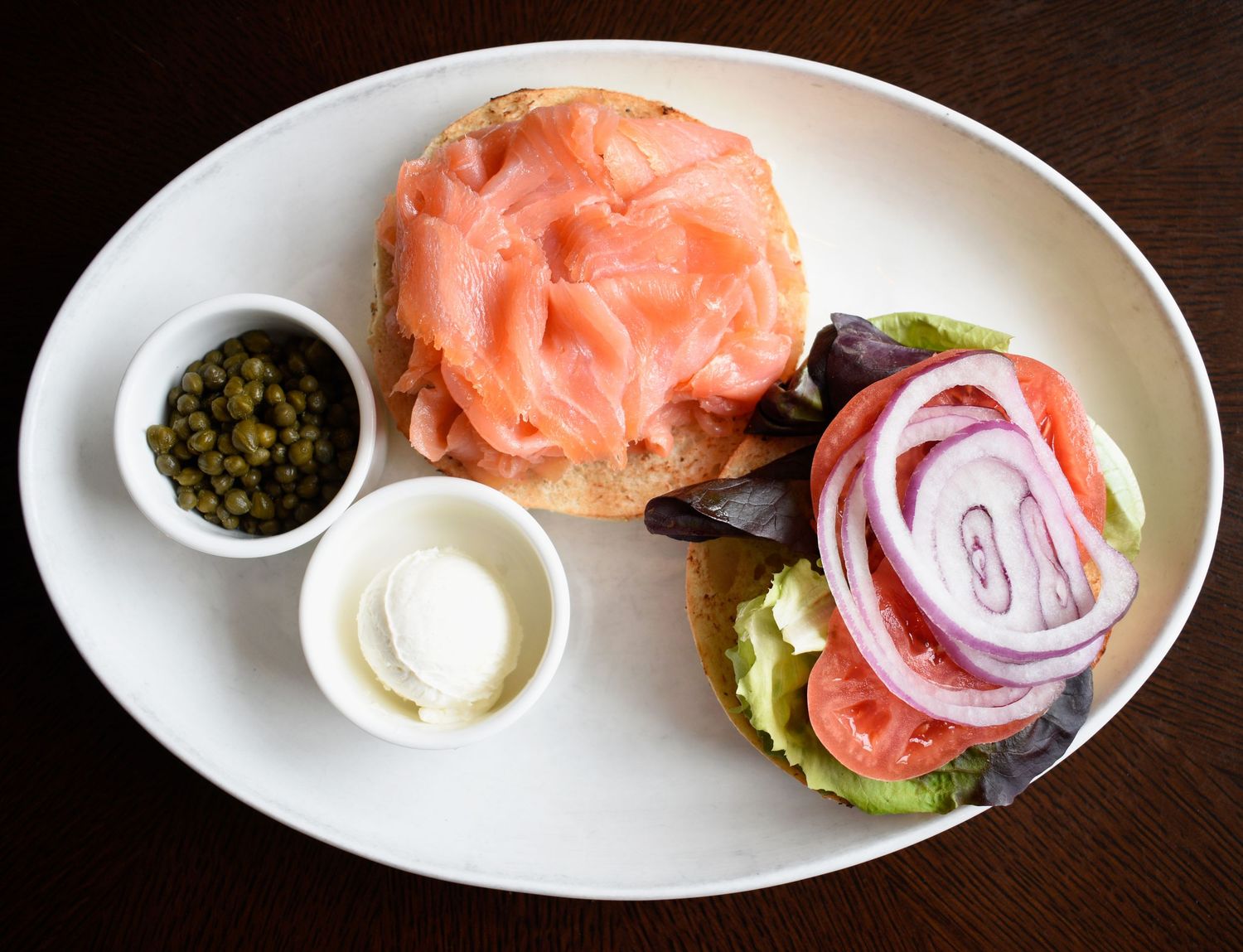 Smoked salmon bagel with capers, cream cheese, red onion, avocado, and mixed greens on a white plate.