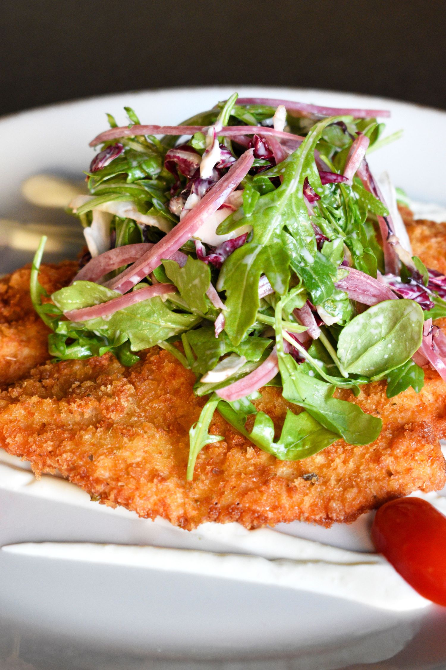 Crispy breaded chicken schnitzel topped with fresh arugula and red onion salad on a white plate.