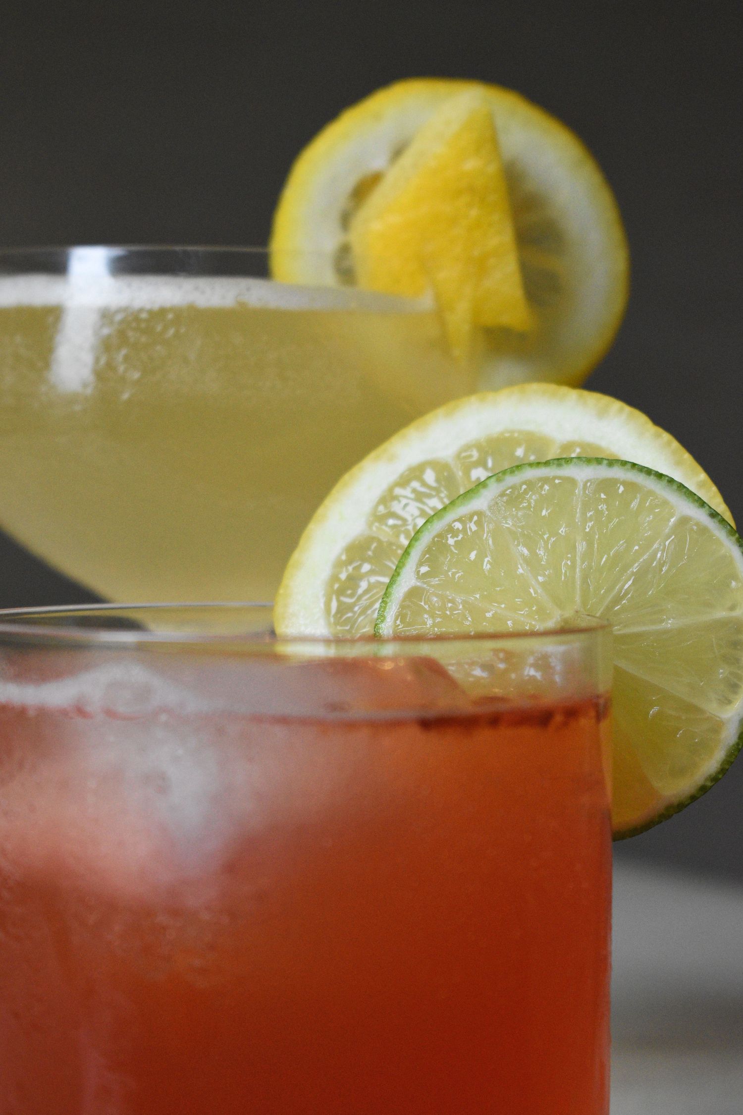 Close-up of a red cocktail with lemon and lime garnish beside a yellow coupe cocktail on a gray background.