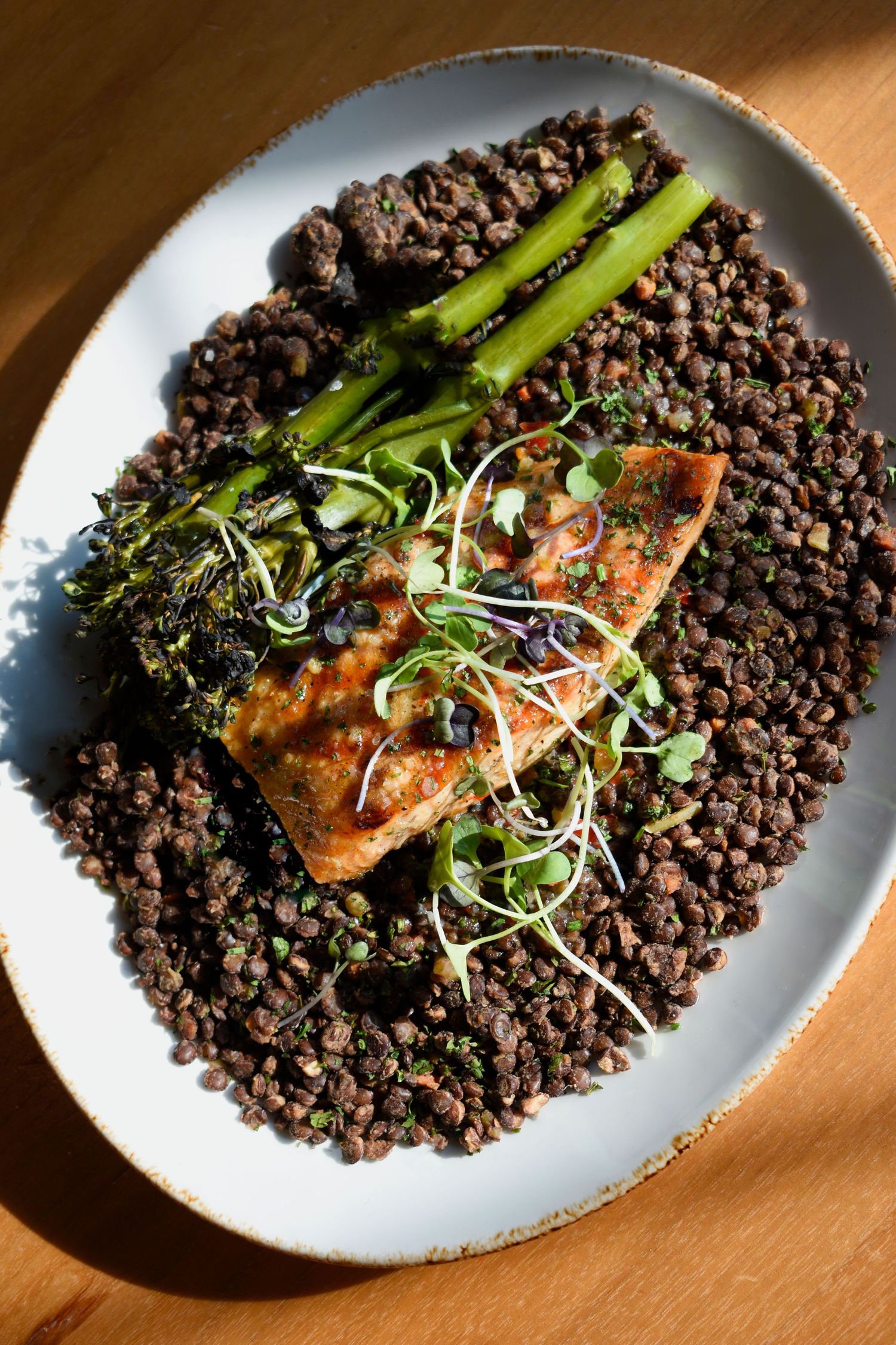 Seared salmon fillet served on black beluga lentils with roasted broccolini and fresh microgreens on a white plate.