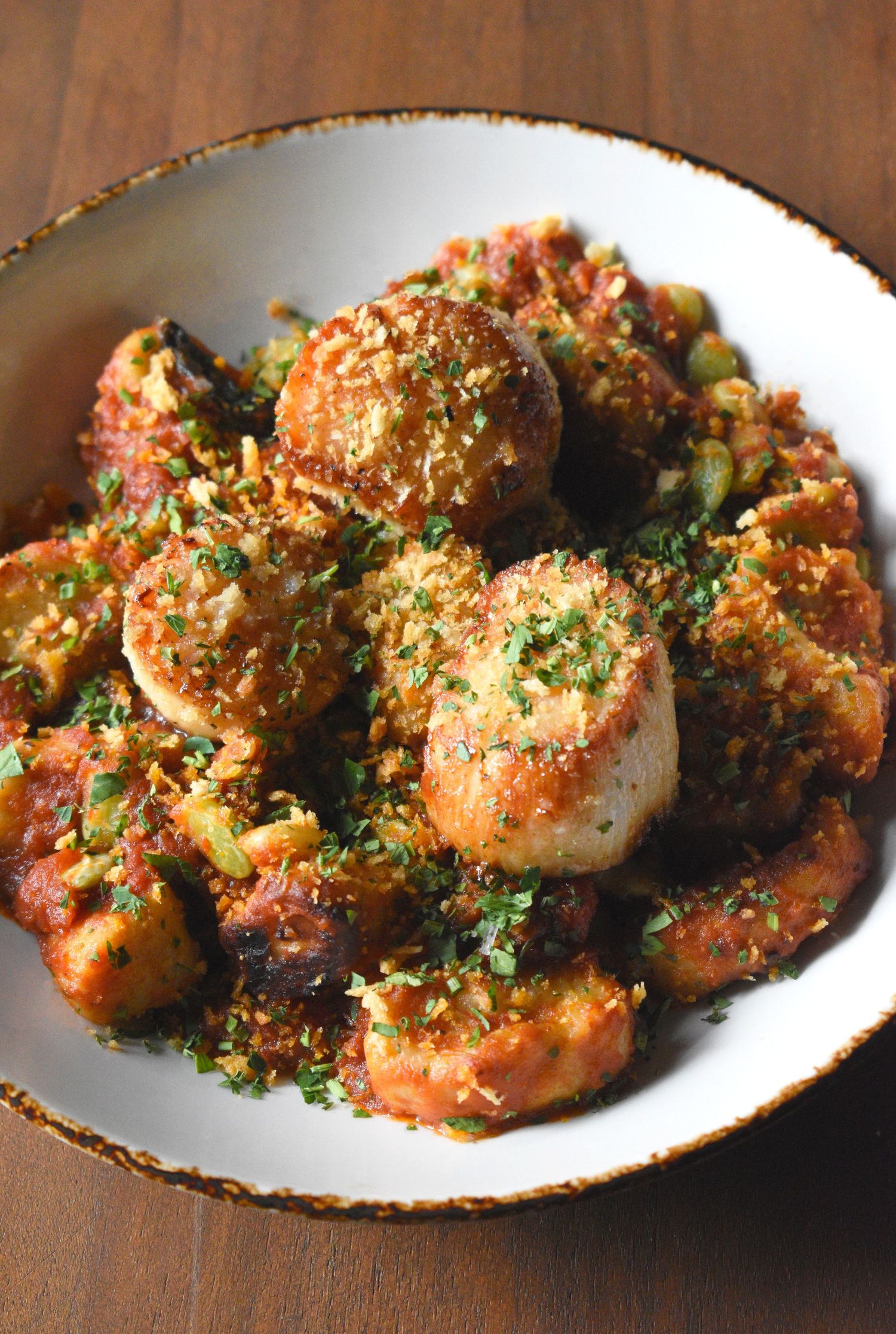 Bowl of seared scallops and shrimp in rich tomato sauce topped with fresh herbs and breadcrumbs.