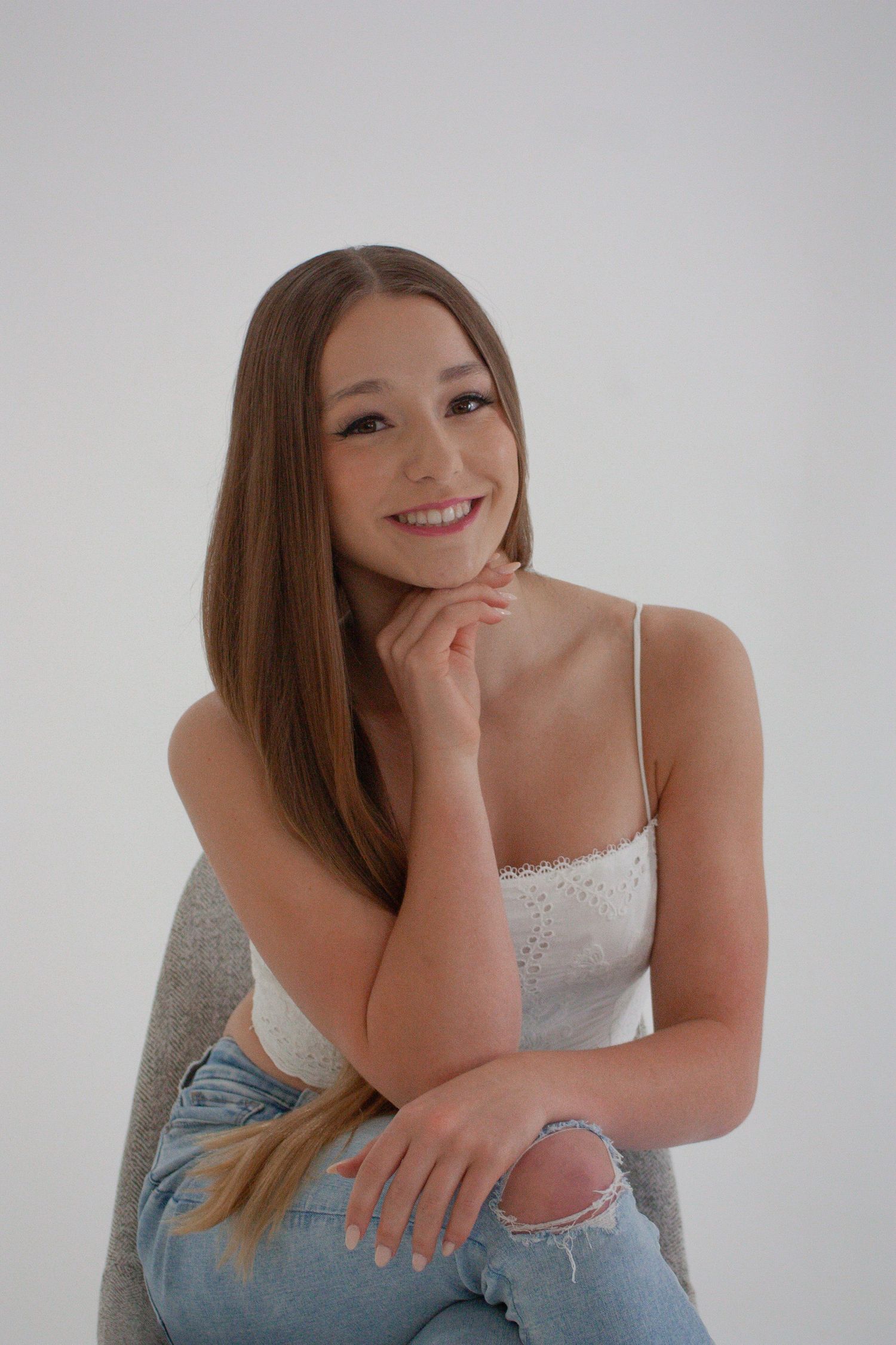 Young woman with long brown hair smiling warmly, wearing white crop top and ripped jeans in a bright studio.