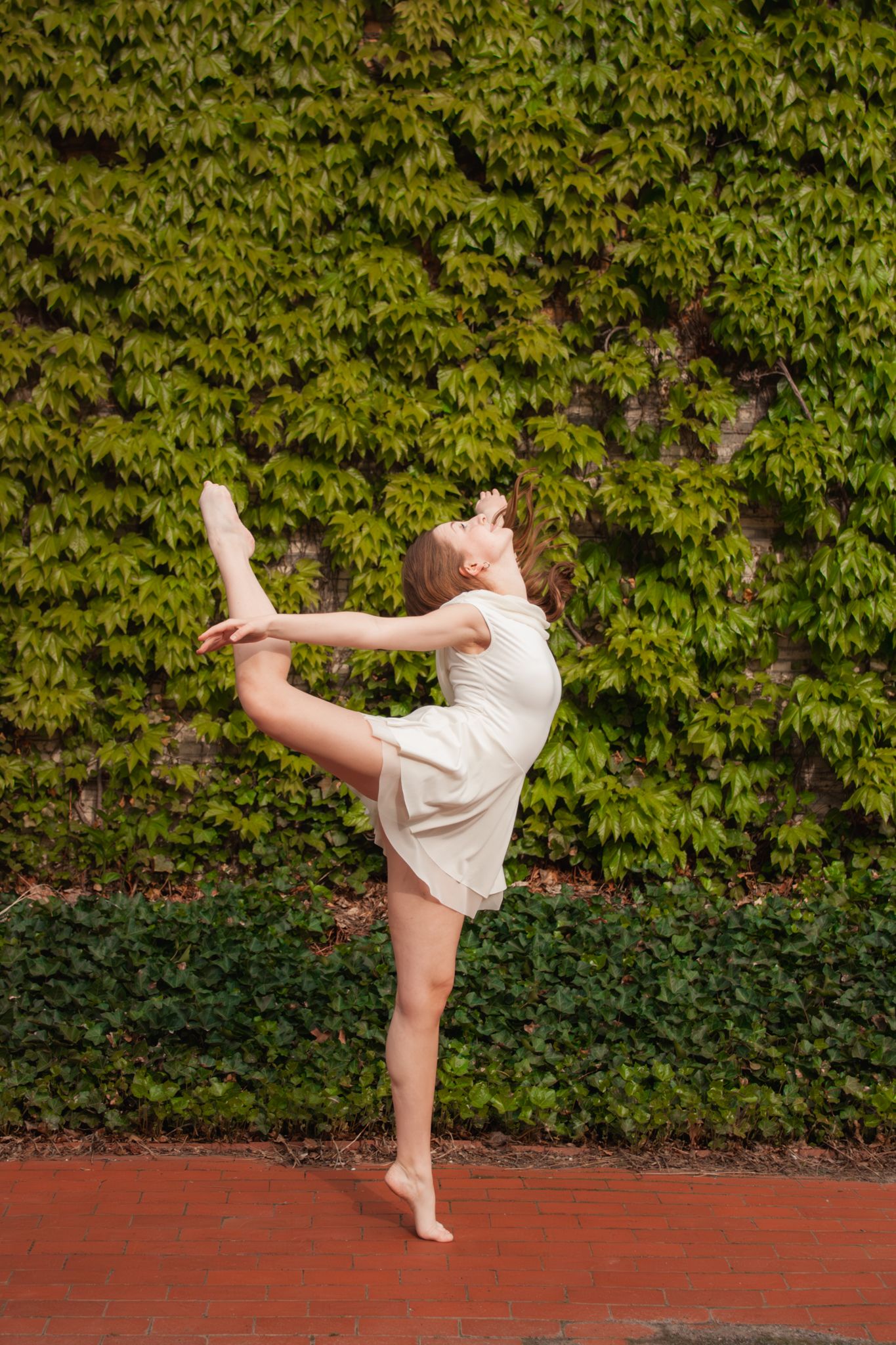 A barefoot dancer in a white dress performs a standing back bend while holding her leg up outdoors.