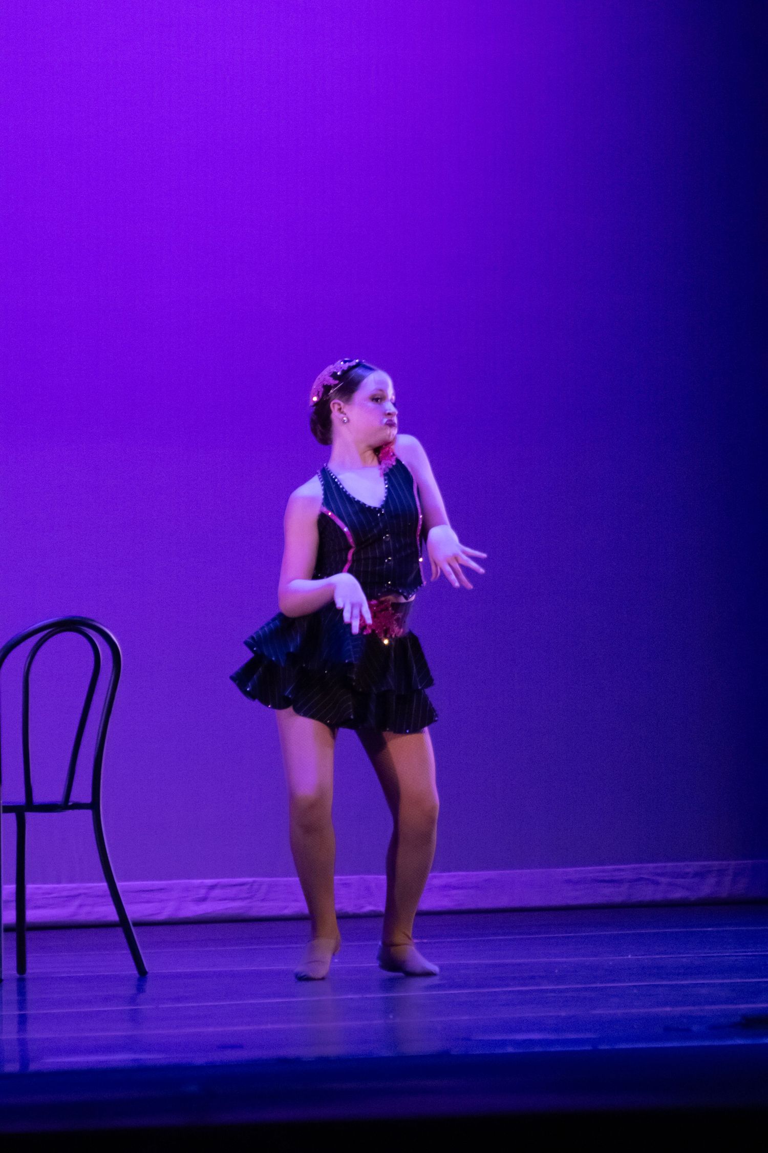 A young dancer in a black ruffled dress performs on stage beside a decorative chair under purple light.