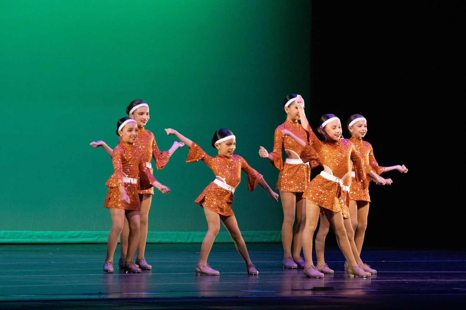 Six young dancers in matching orange sequined costumes perform a synchronized routine on a green-lit stage.