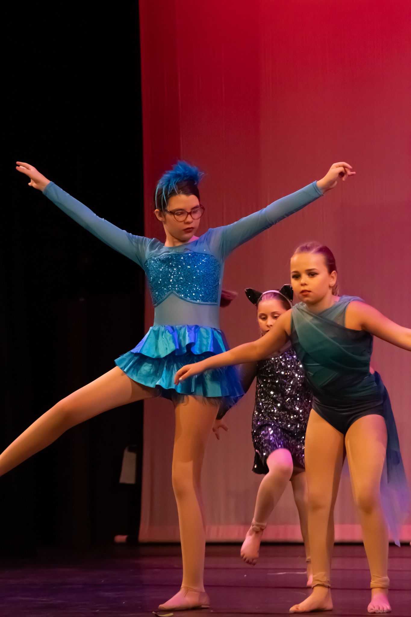 Three young dancers in blue and teal costumes perform on a red-lit stage.