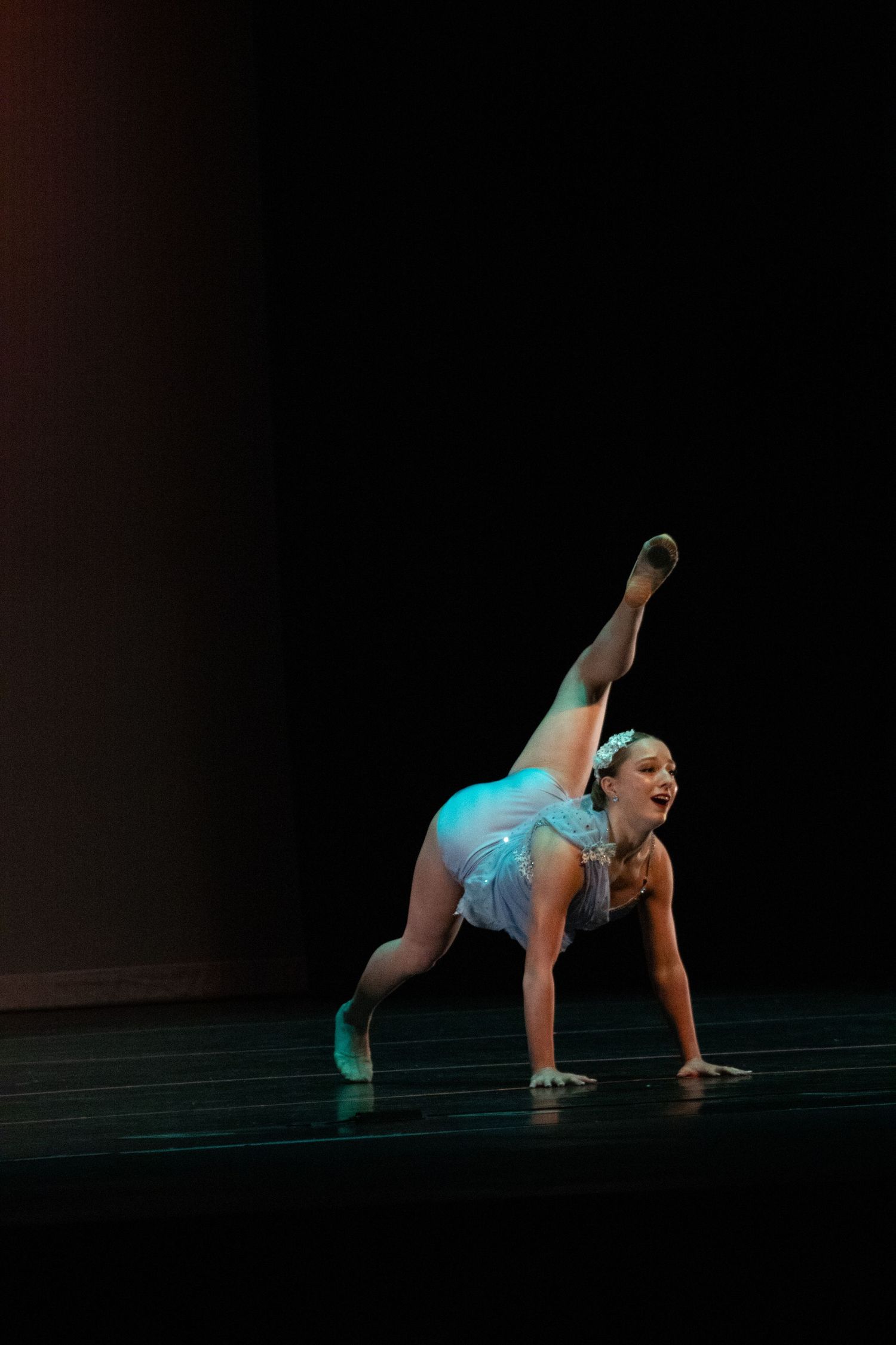 A smiling dancer in light blue costume performs a floor acrobatic pose on a dark stage.