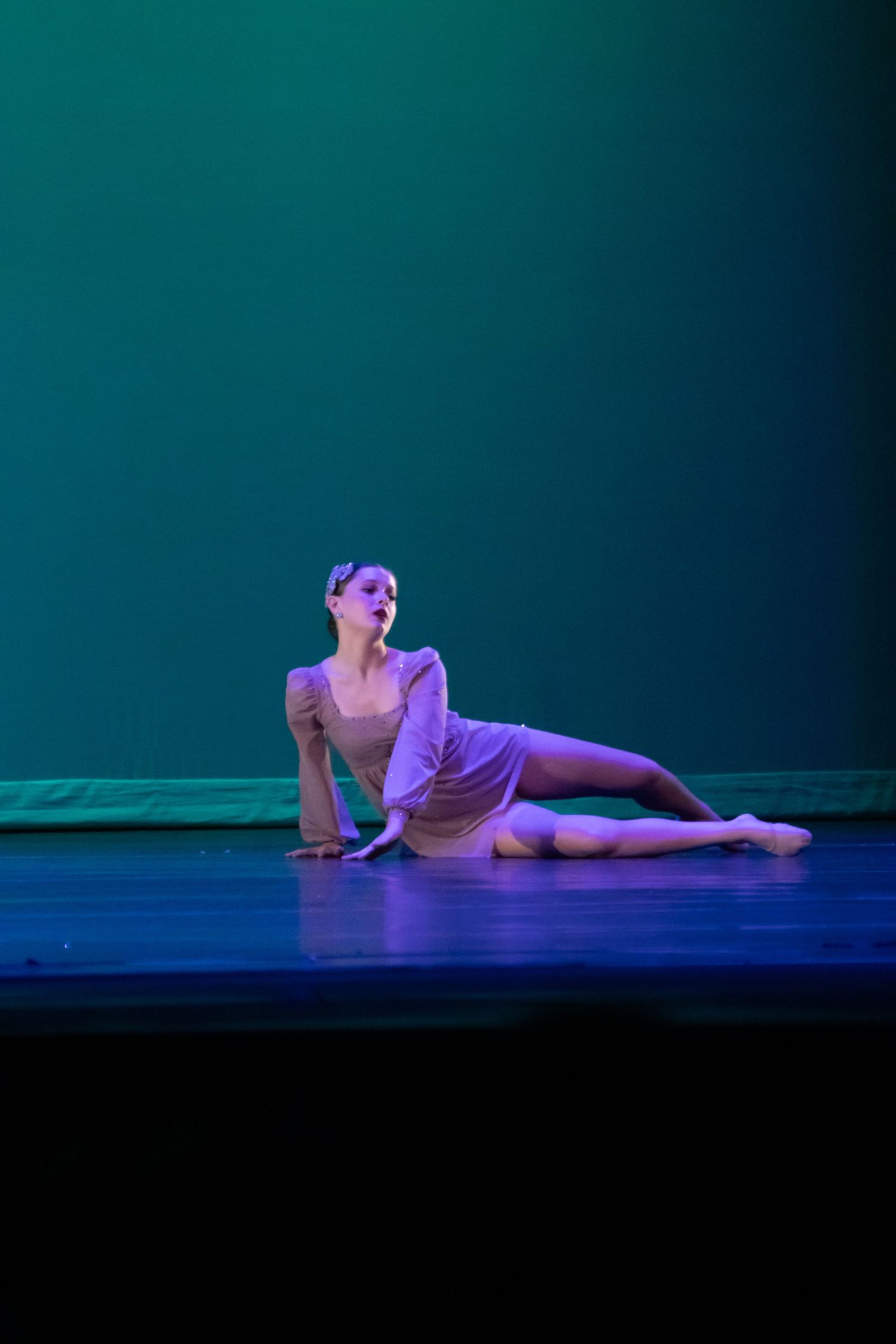 A dancer in a lavender dress poses gracefully on the floor under teal and blue stage lighting.