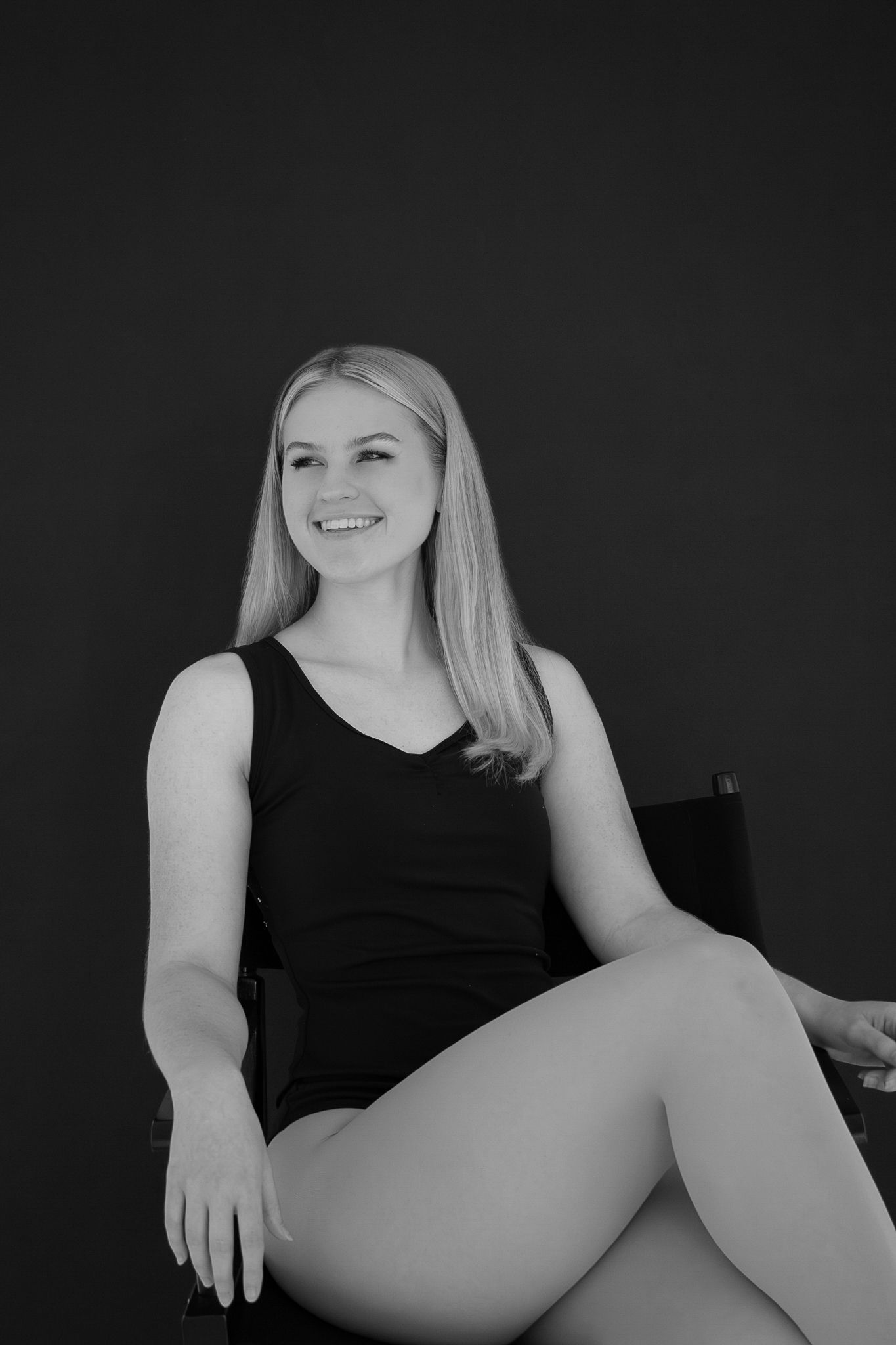 A smiling blonde woman sits relaxed in a chair against a dark background in black and white.