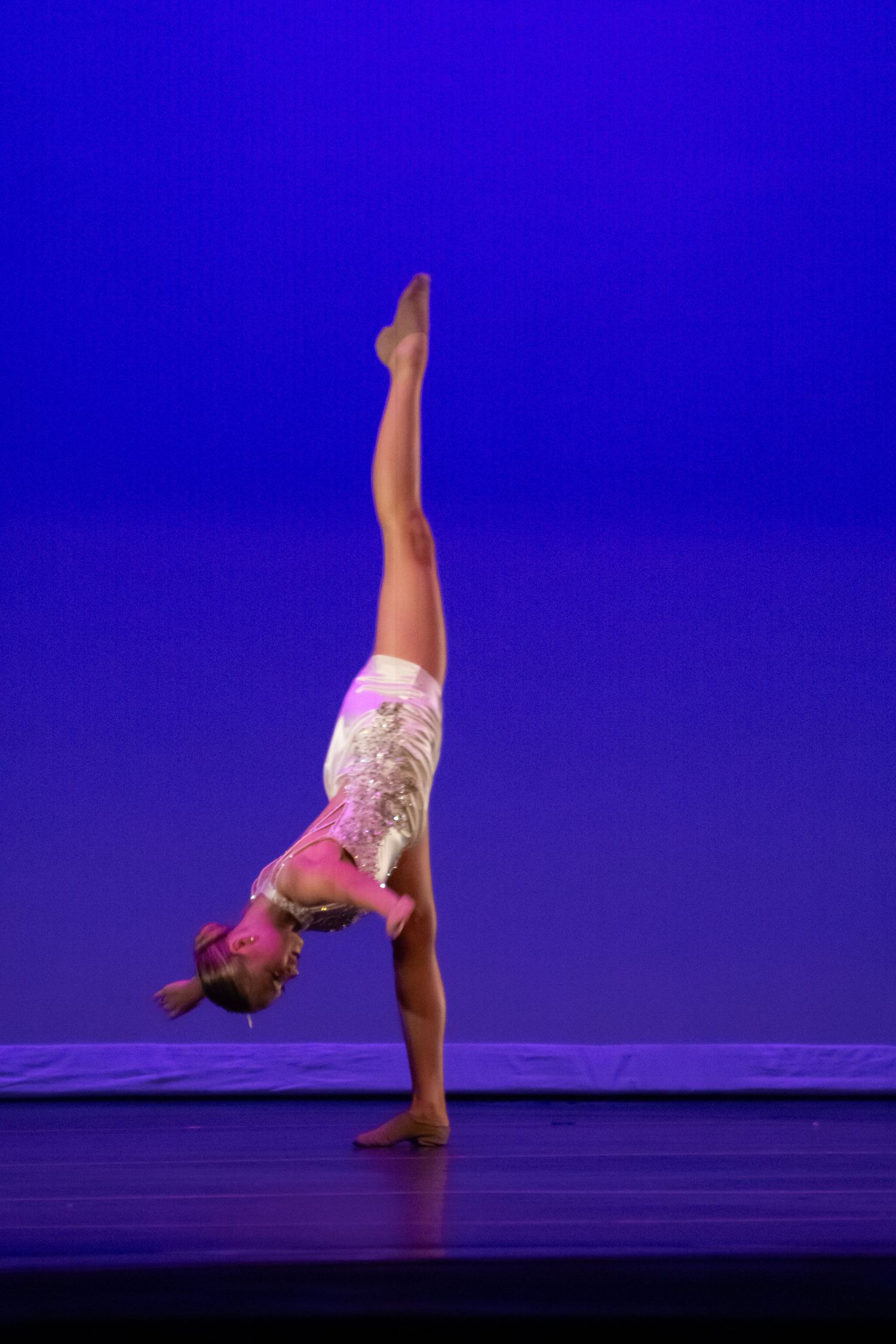 A dancer in a pink costume performs a handstand with legs split vertically on a blue-lit stage.